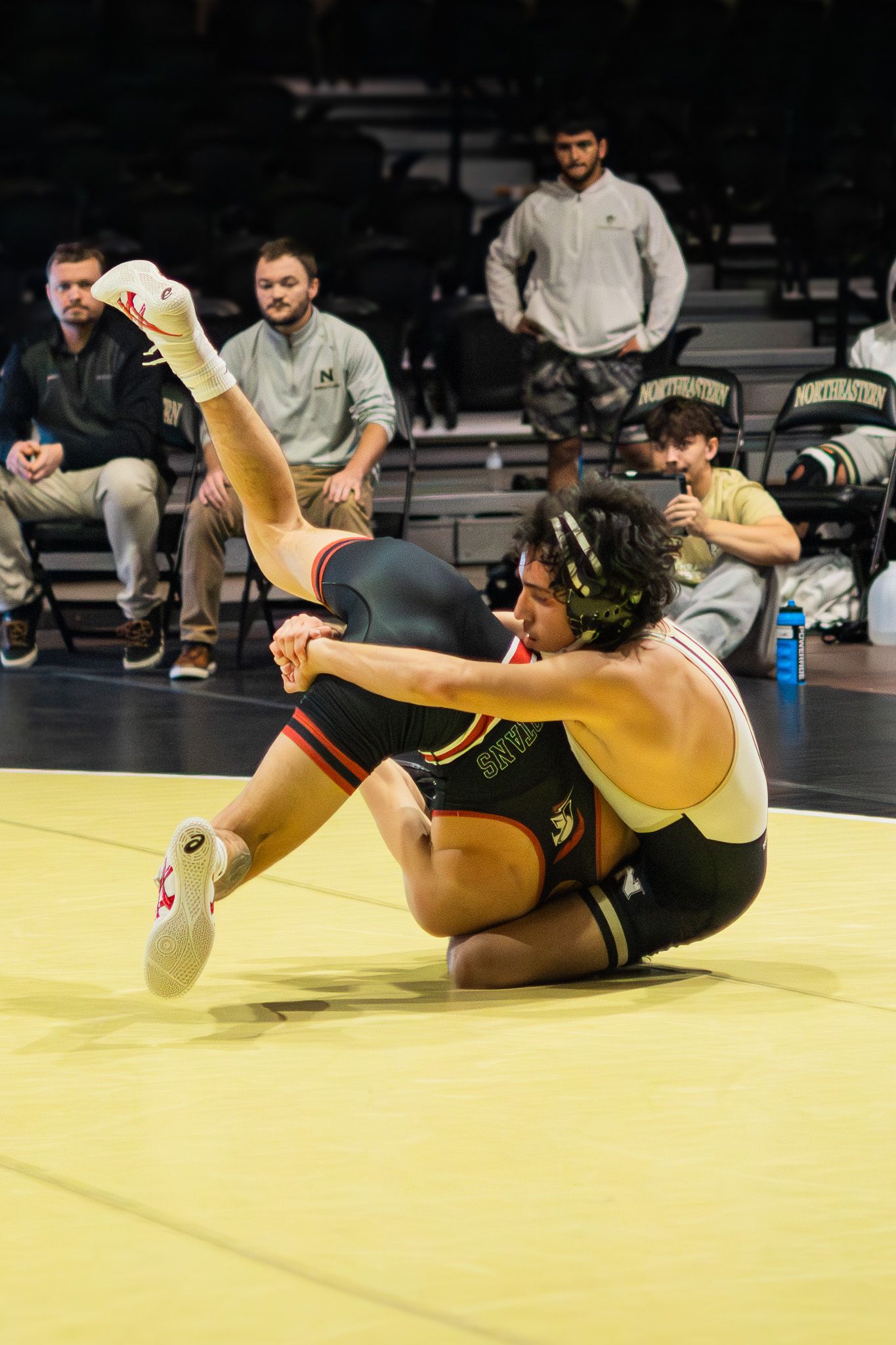 Wrestler in gold singlet executes a takedown move on an opponent during an indoor wrestling competition.