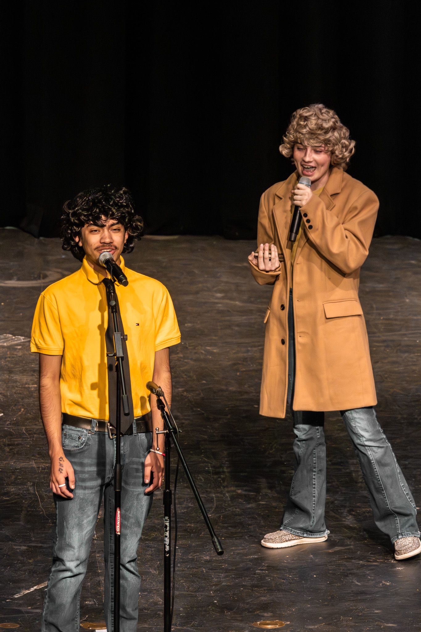 Two young male performers on stage, one in yellow shirt and one in tan coat, during live show.