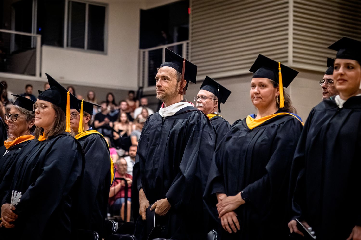Row of graduates in black caps and gowns with gold honor cords stand at Northeastern Junior College ceremony.