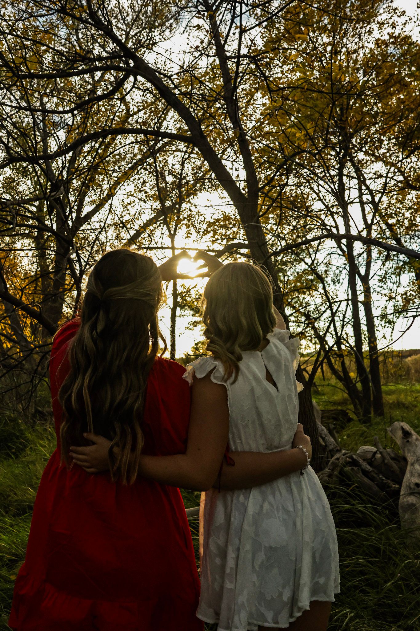 Two girls in red and white dresses embrace while watching a golden sunset through autumn trees.