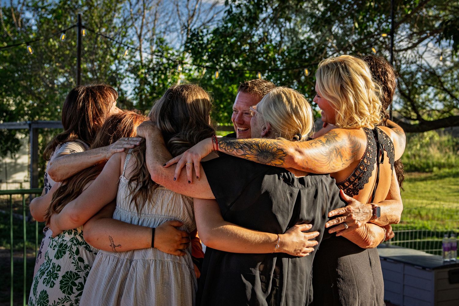 A group of women share a joyful group hug outdoors surrounded by green trees at a backyard gathering.