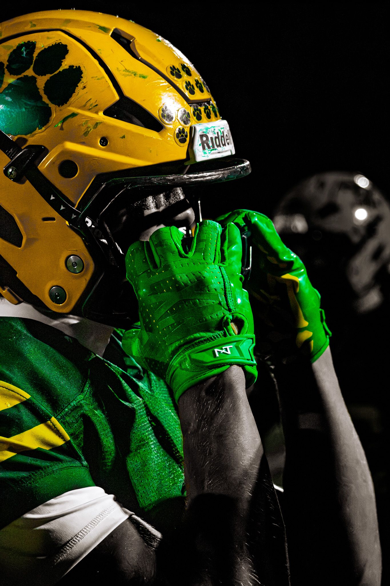 High school football player in green and gold uniform and Riddell helmet under dramatic dark lighting.