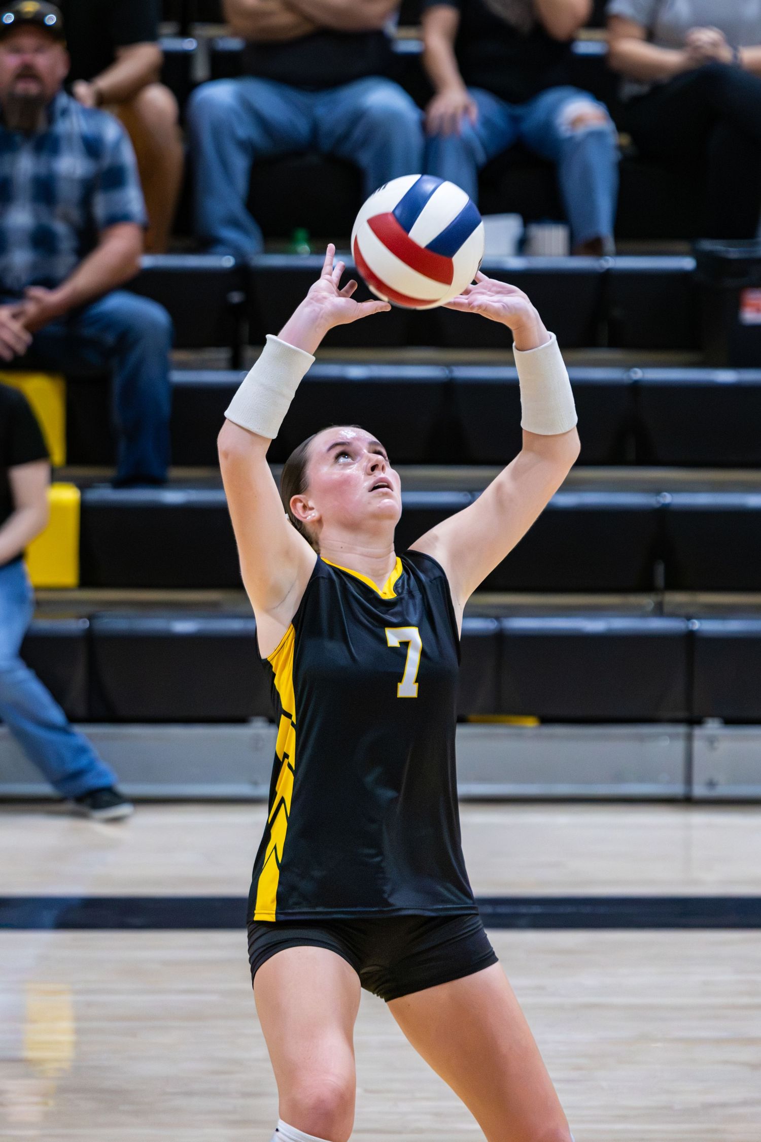 Volleyball player number 7 in black and gold uniform sets the ball during a high school match.