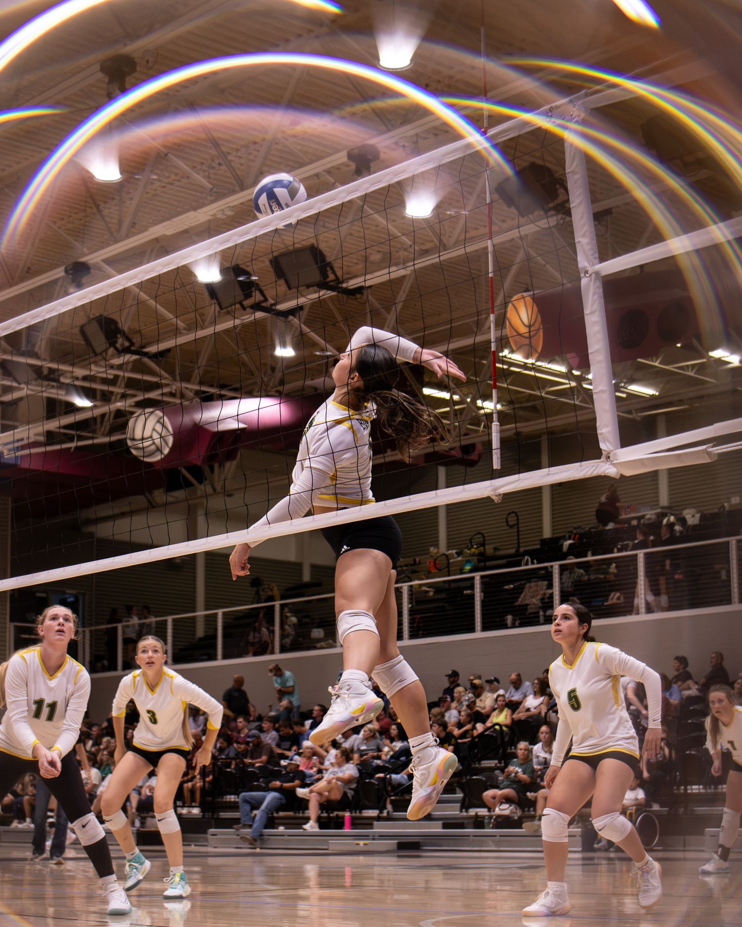 Volleyball player leaps to spike the ball as teammates watch during an indoor high school match with packed stands.