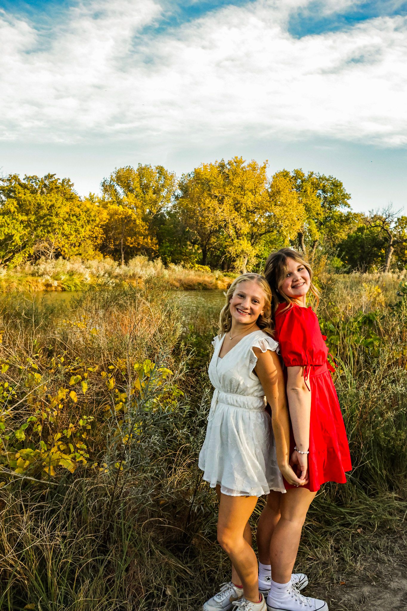 Two smiling girls in white and red dresses pose back-to-back in a sunny autumn meadow.