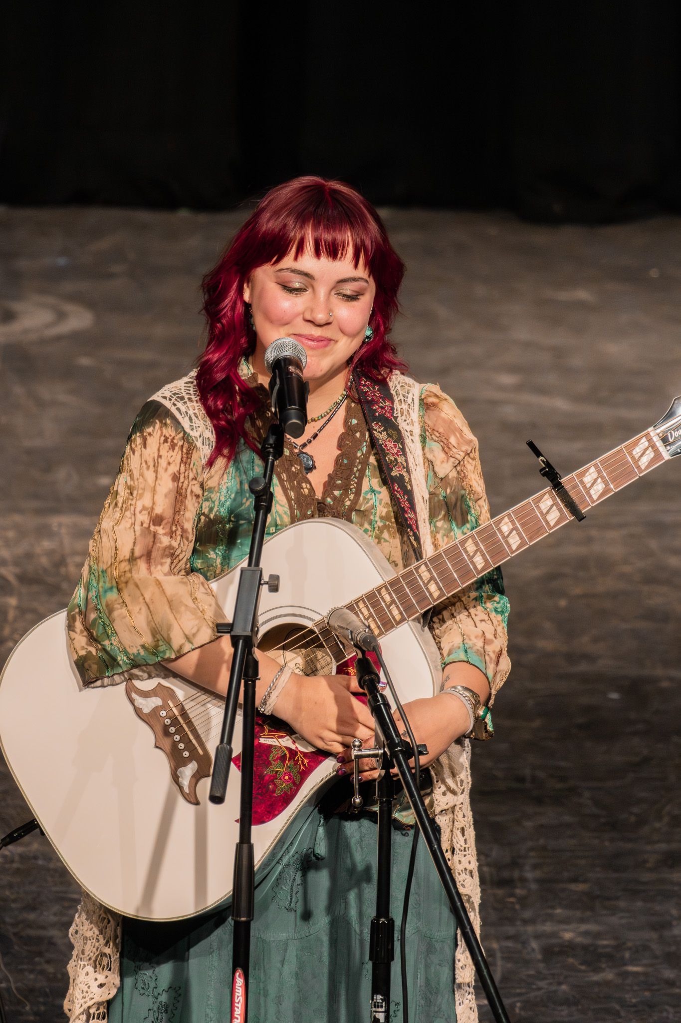 Red-haired female musician smiling while playing white acoustic guitar on stage in bohemian floral outfit.