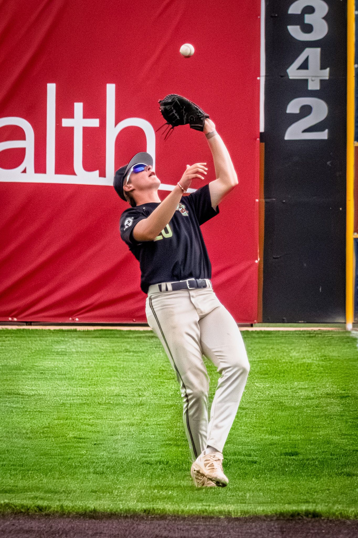 A baseball outfielder in navy uniform catches a fly ball near the 342-foot warning track marker.