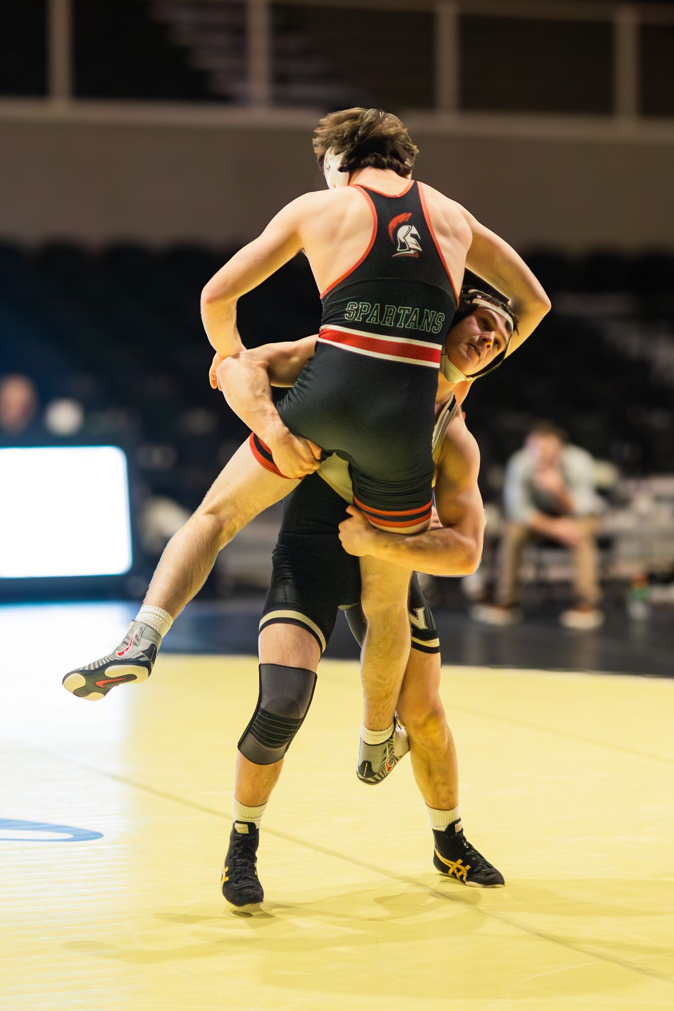 Wrestler in black Spartans singlet lifts opponent off mat during college wrestling match.