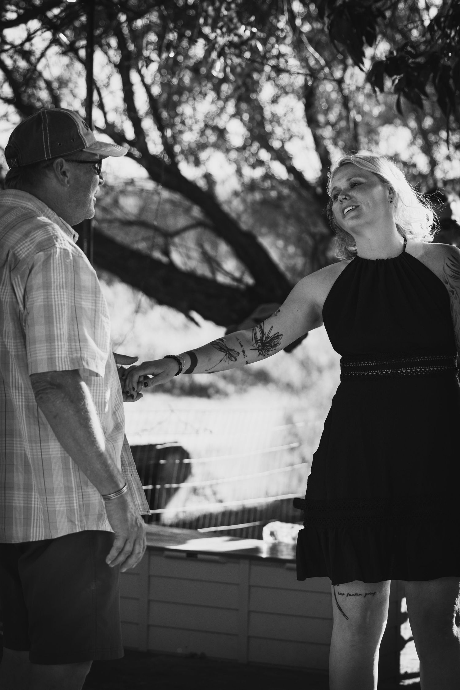 Black and white photo of an older man holding hands with a tattooed woman in a black dress outdoors.