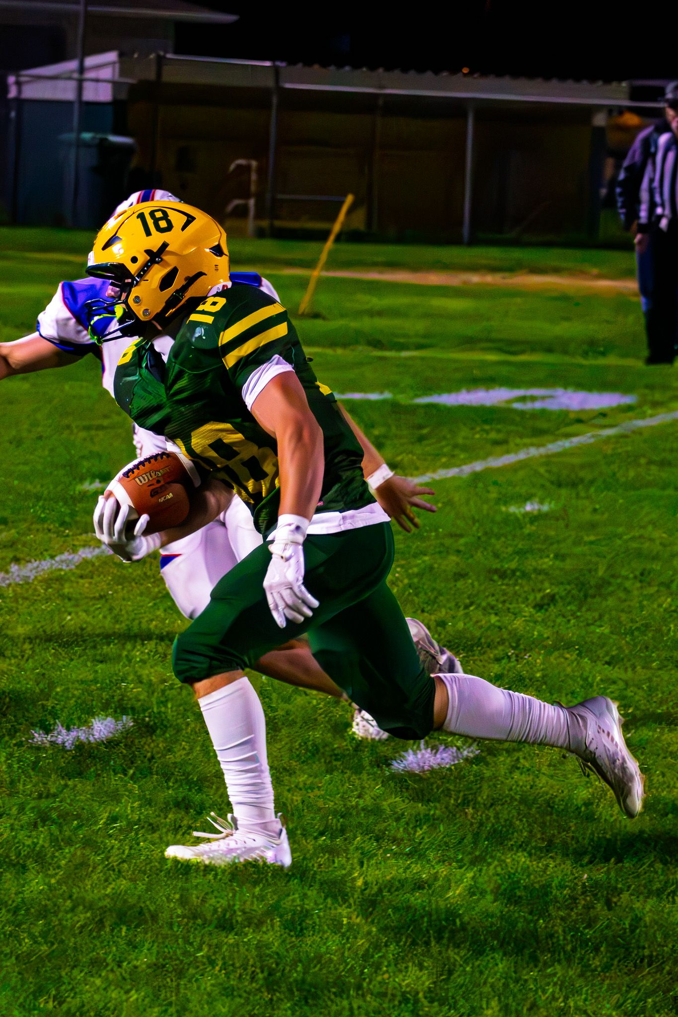Green and gold football player #18 carries the ball while being tackled during a night game.