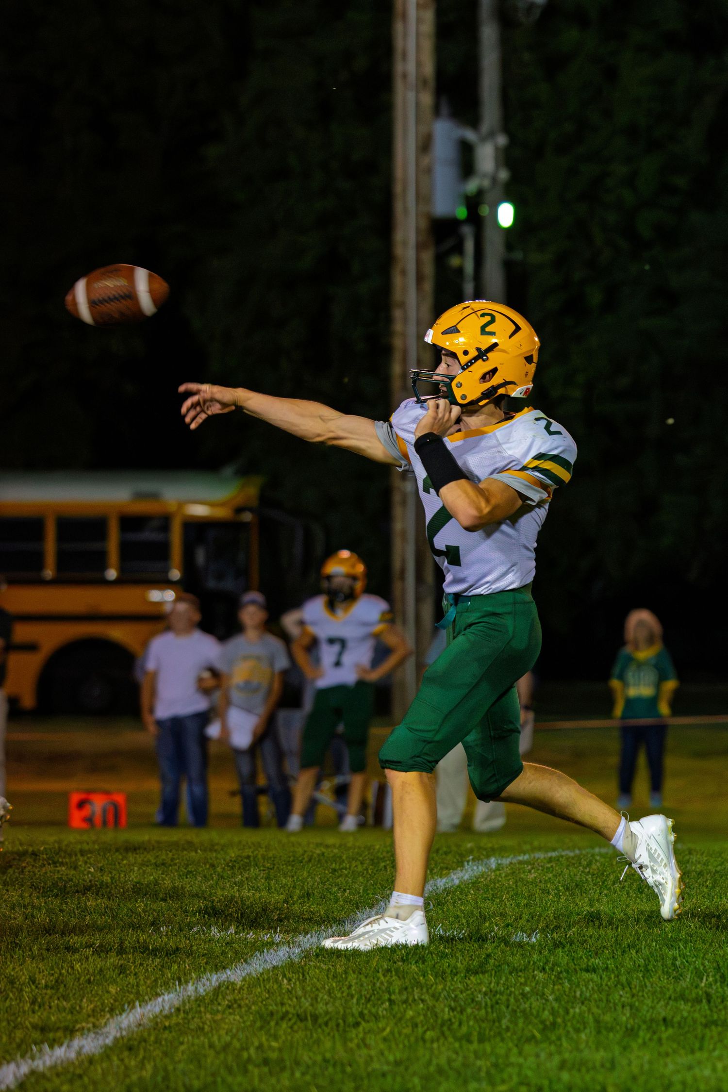 Youth football player #2 in green and gold uniform punts the ball near the sideline during a night game.