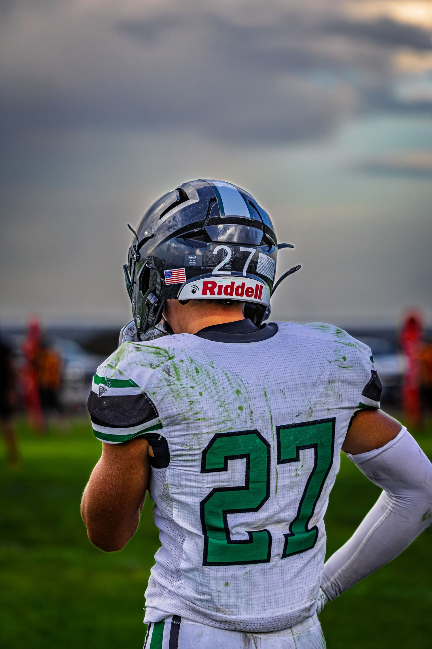 Back view of football player #27 in white Riddell helmet and green jersey on field under cloudy sky.