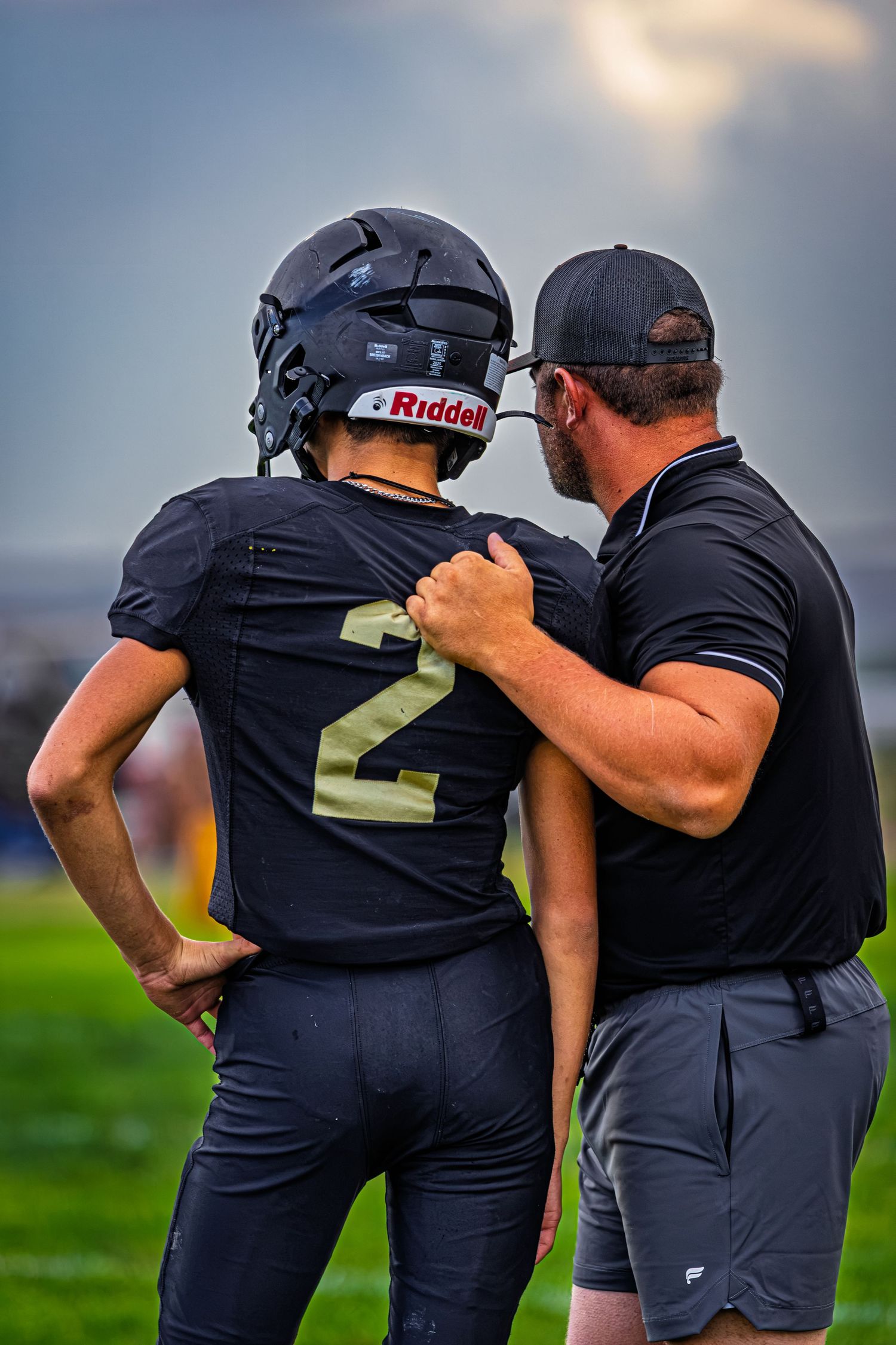 A football coach in a black polo gives sideline advice to player number 2 wearing a Riddell helmet.