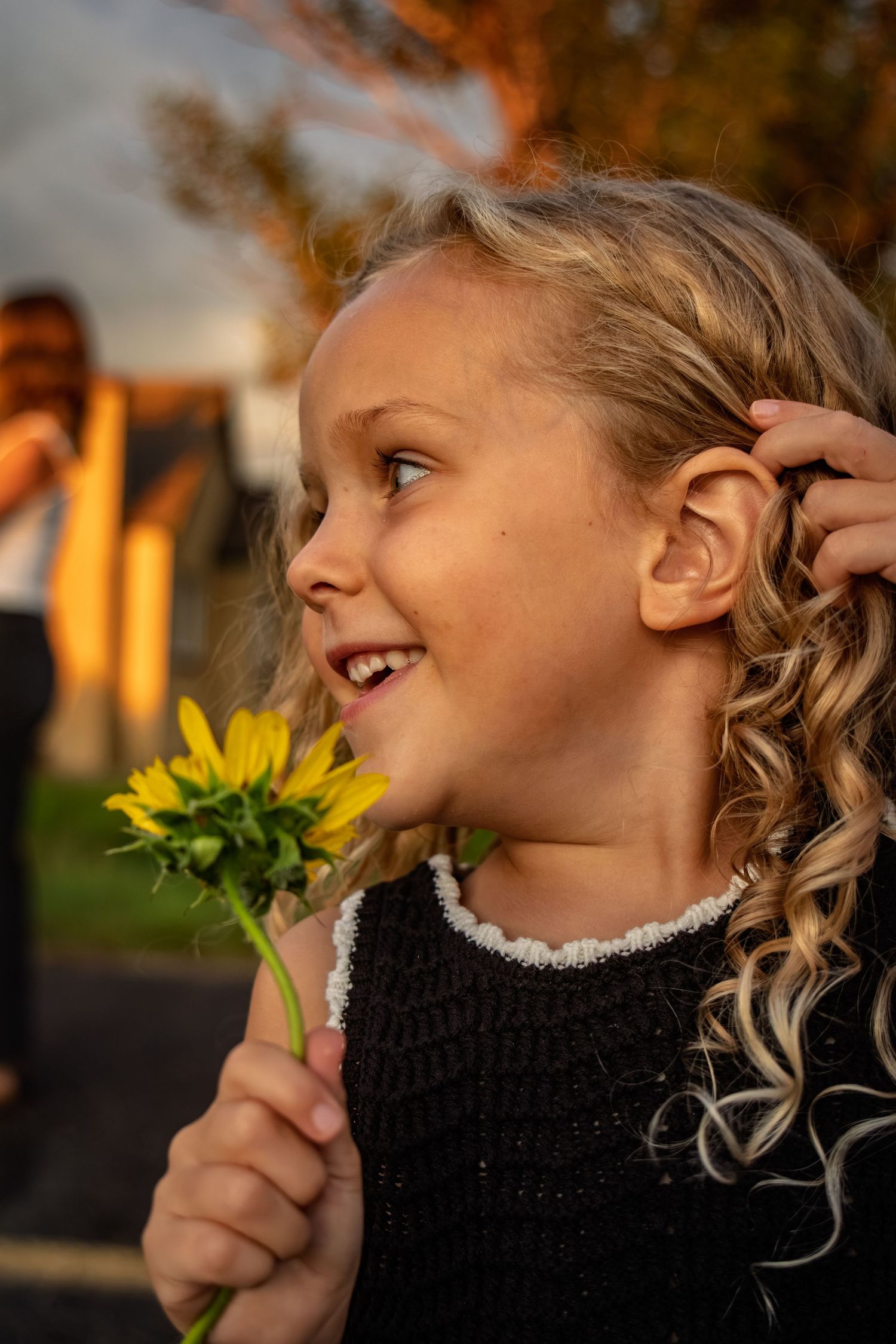 Happy curly-haired blonde girl smiling while holding a yellow sunflower in golden sunset light.