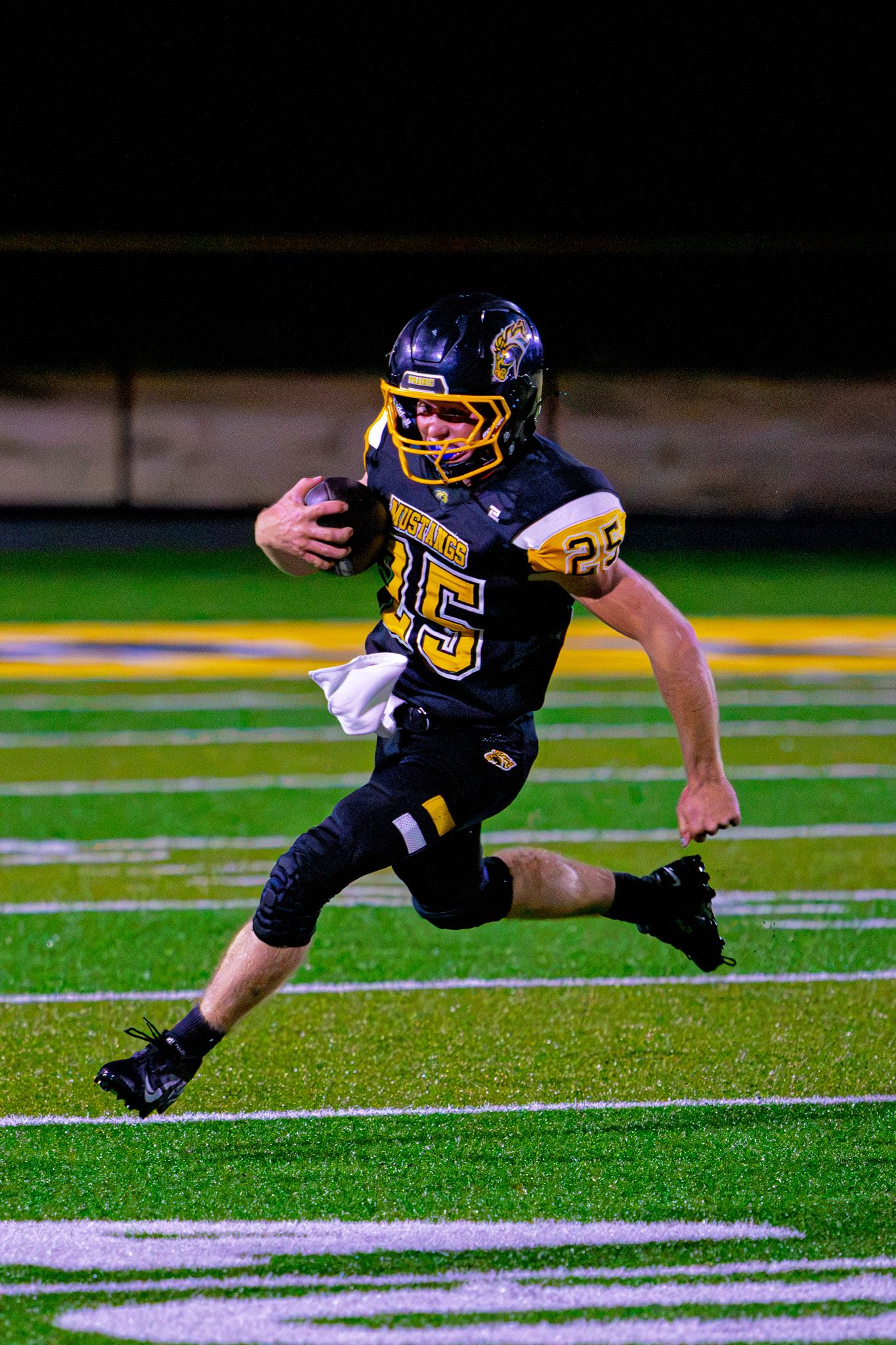 High school football player number 25 in black and gold uniform runs with the ball during a night game.