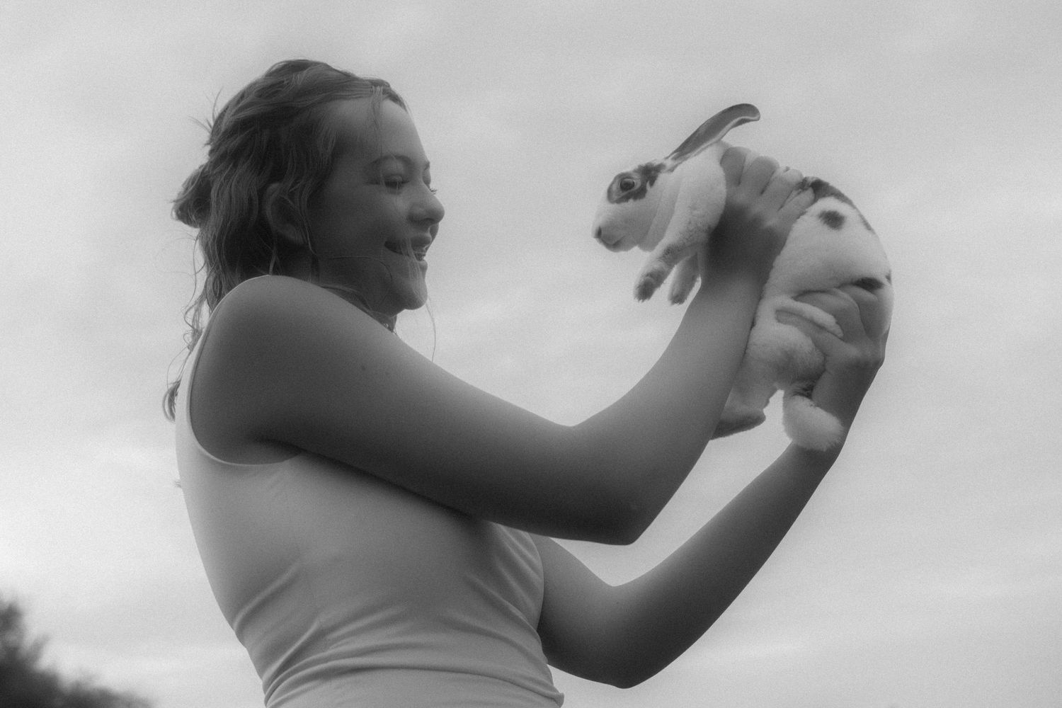 Black and white photo of a smiling young girl holding a rabbit up against a cloudy sky.