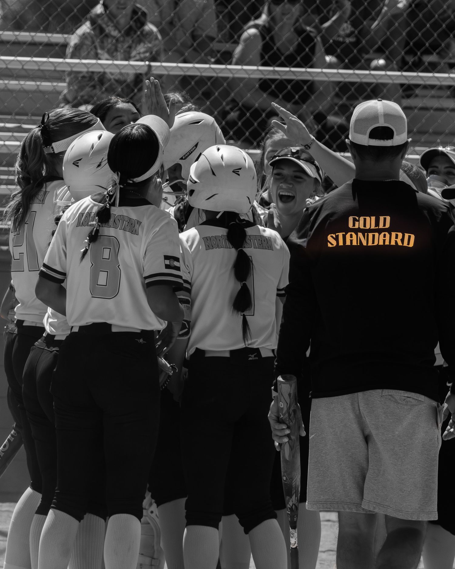 Black and white photo of softball team huddle with Gold Standard coach jersey visible.