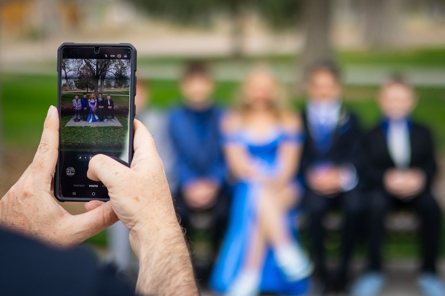 Person photographing a group of teens in formal prom attire seated on a bench outdoors.