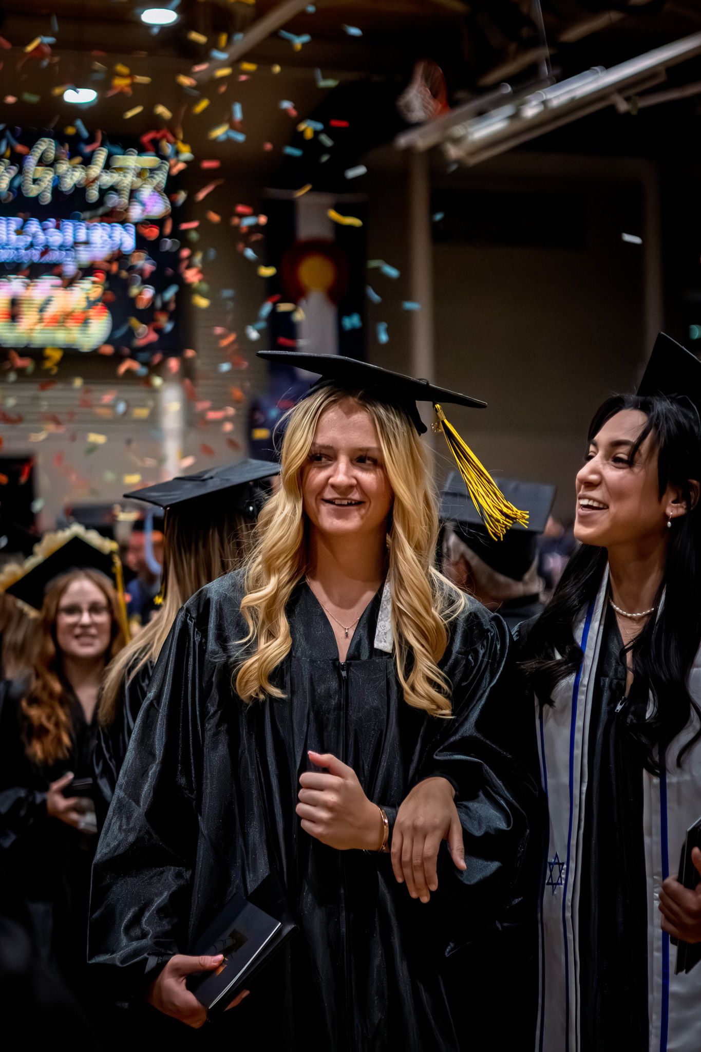 Two smiling graduates in black caps and gowns celebrate amid colorful confetti at a graduation ceremony.