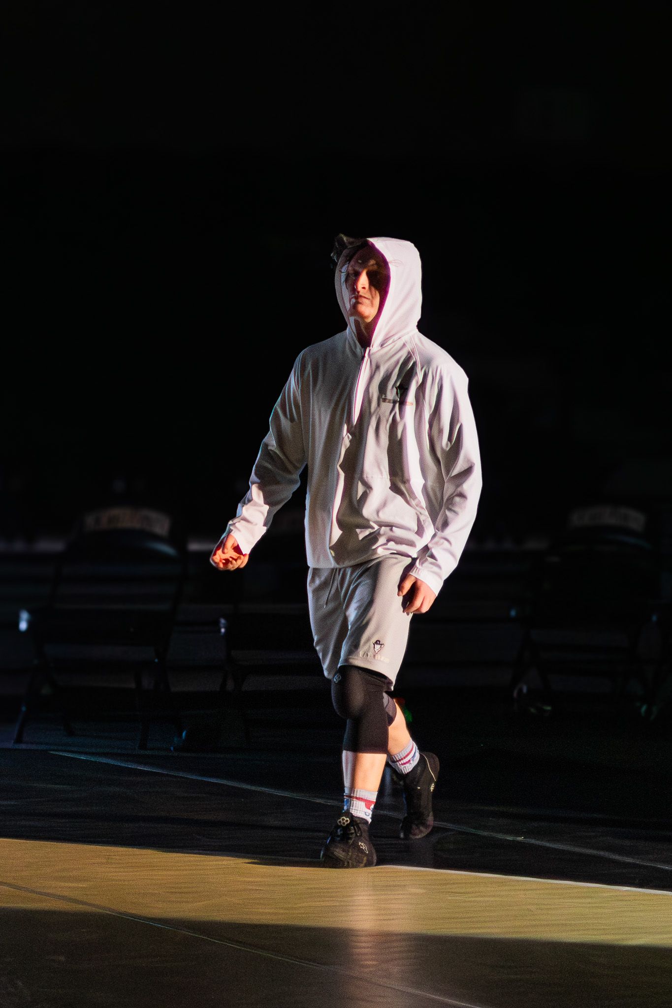 Wrestler in white hoodie and shorts walks dramatically through a dark arena spotlight before a match.