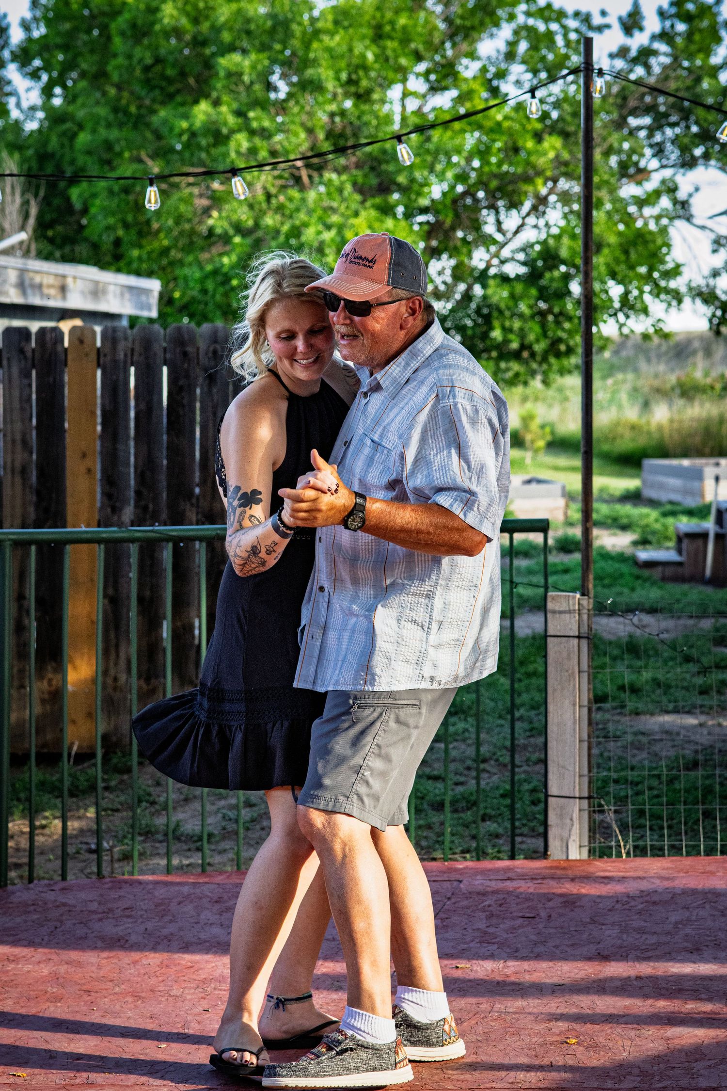 A smiling young woman with tattoos dances with an older man in a cap at an outdoor backyard gathering.