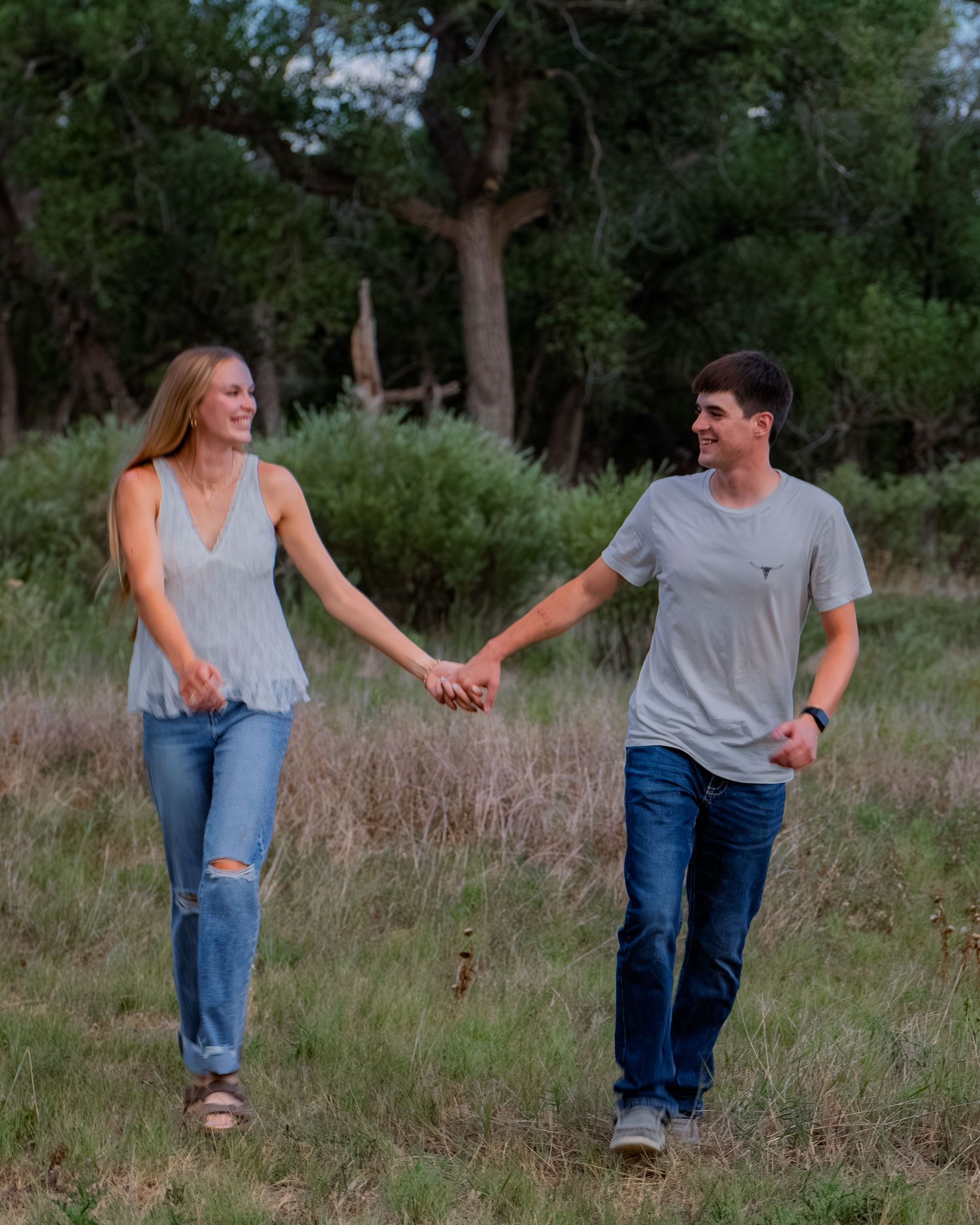 Smiling young couple holding hands while walking through a grassy field with trees behind.
