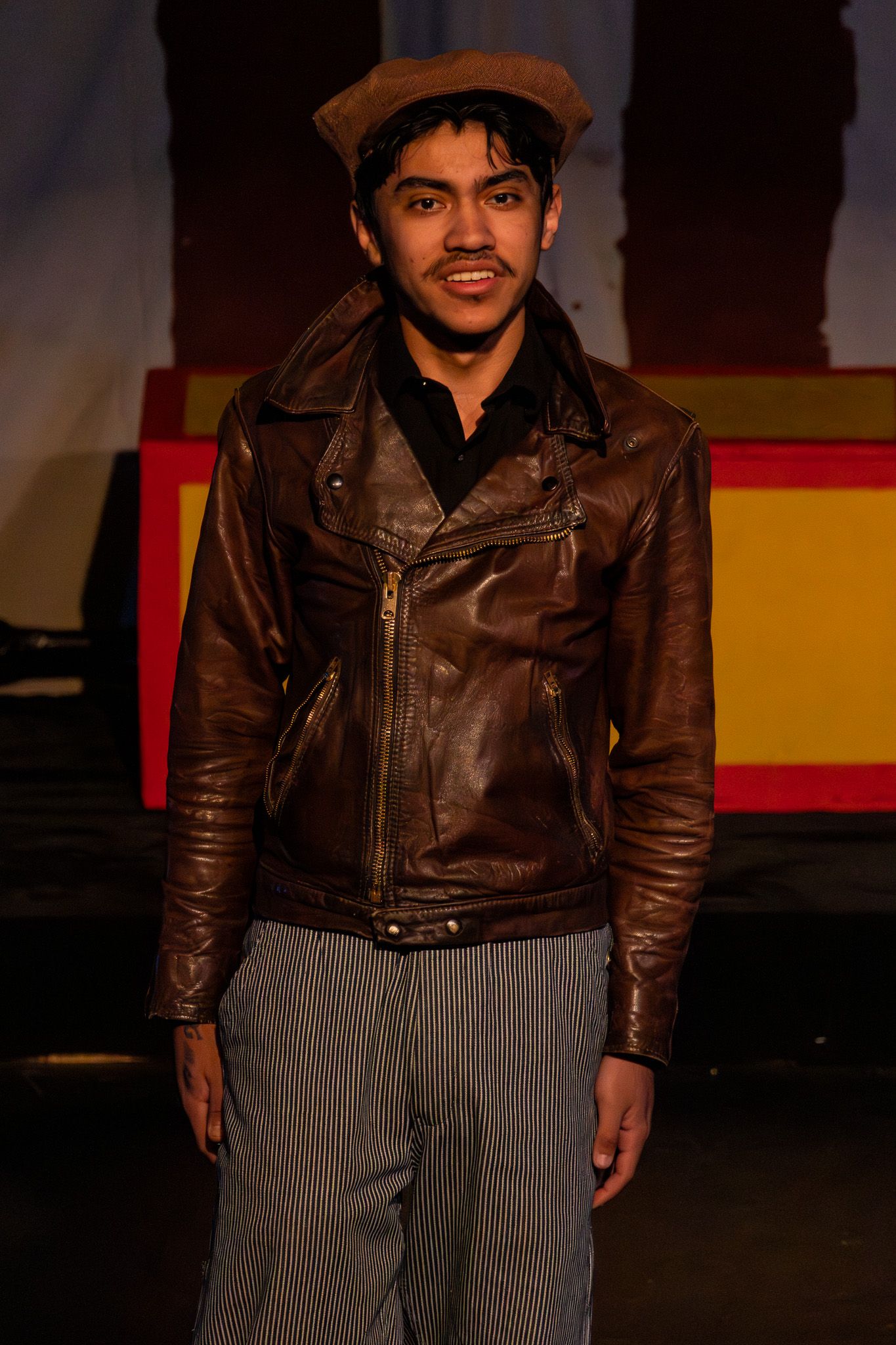 Young male actor smiles on stage wearing a brown leather jacket and hat in a theater production.