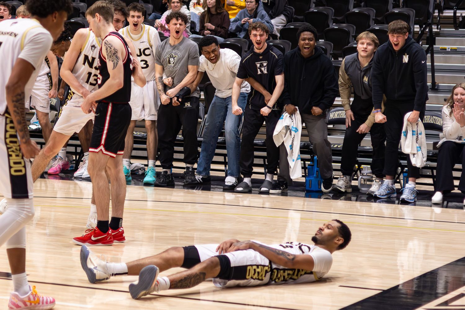 A basketball player lies on the court after a foul as teammates and fans react excitedly on the sideline.