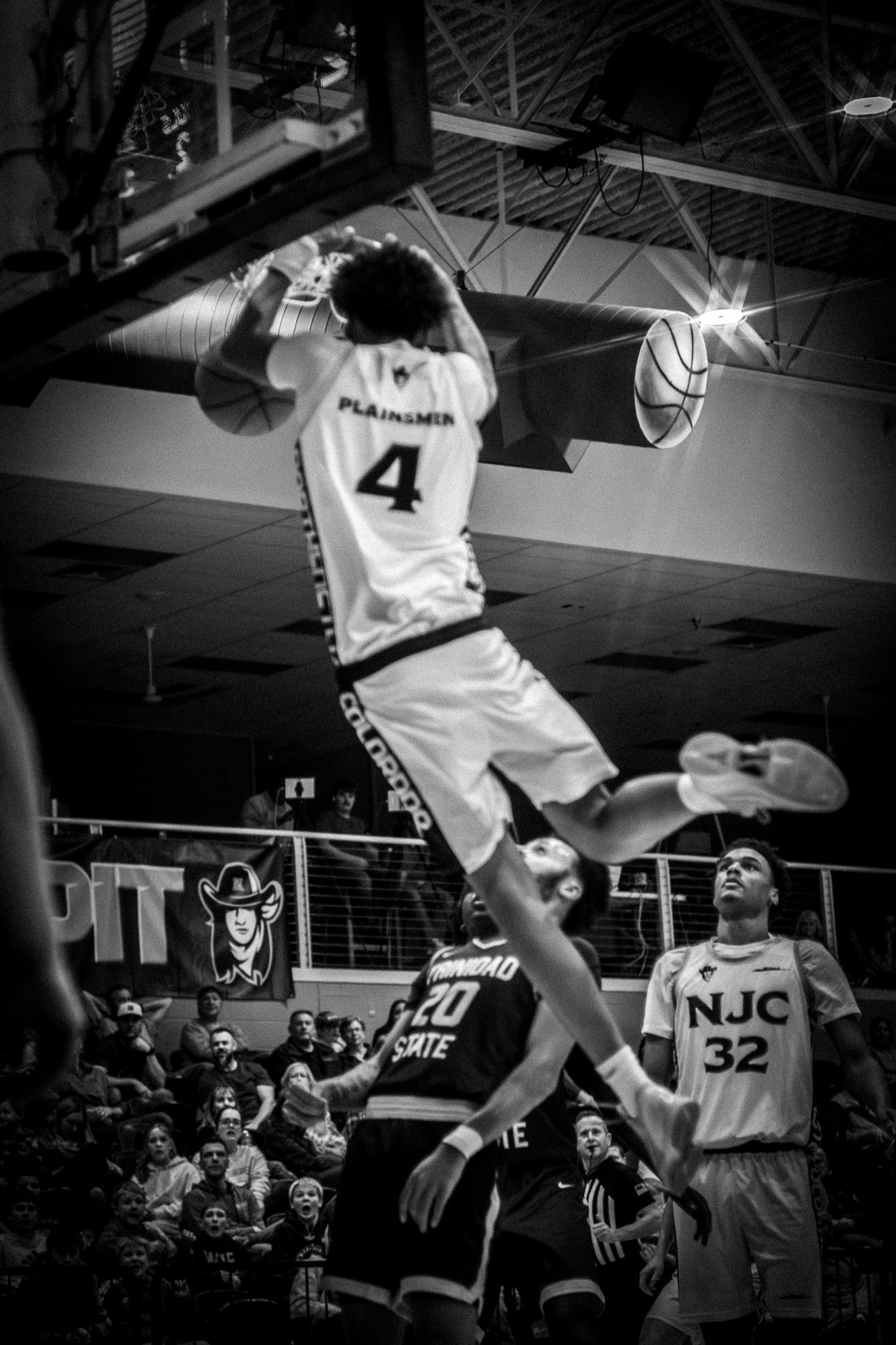 Black and white photo of Plainsmen #4 dunking over a Otero State player in a college basketball game.