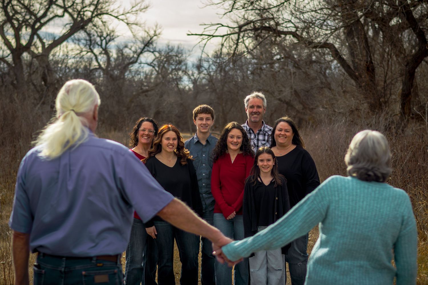 A large multigenerational family holds hands in a circle outdoors among bare winter trees.