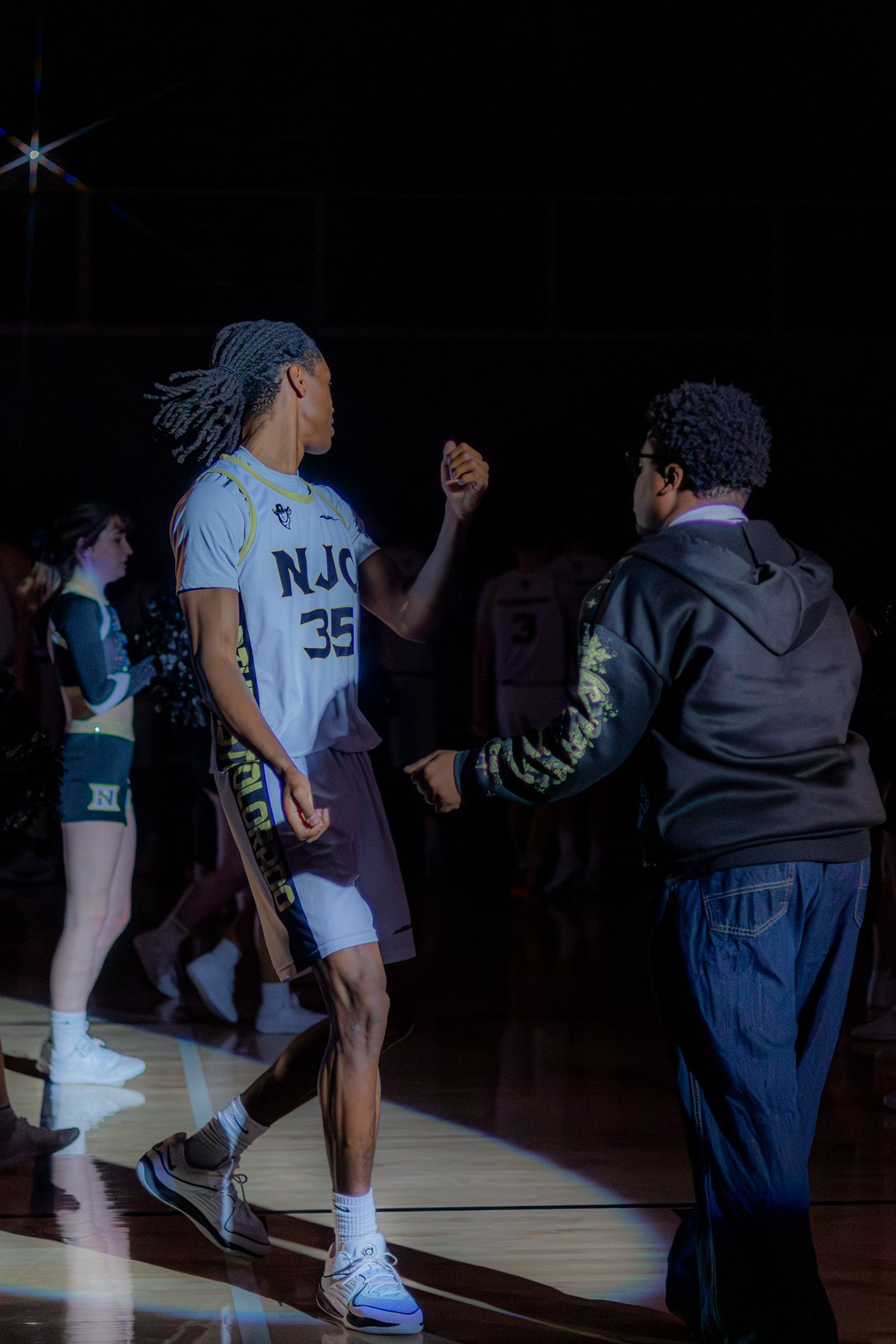 NJC Plainsmen player #35 fist bumps a coach during pre-game introductions in a dark basketball arena.