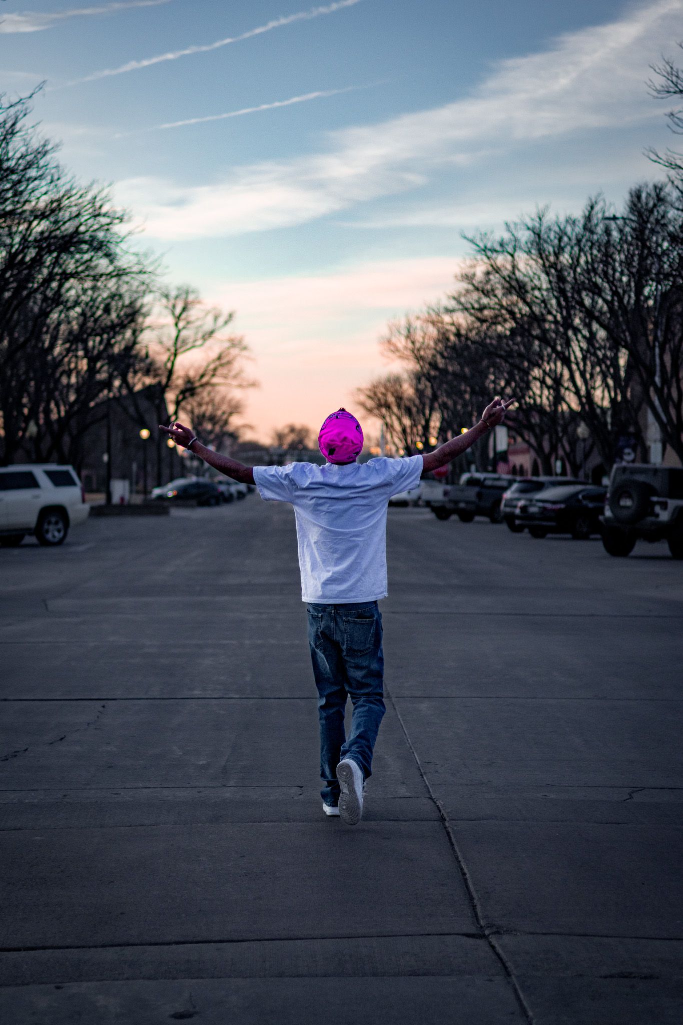 A person in a white tee, jeans, and pink beanie jumps with arms raised on a street at sunset.