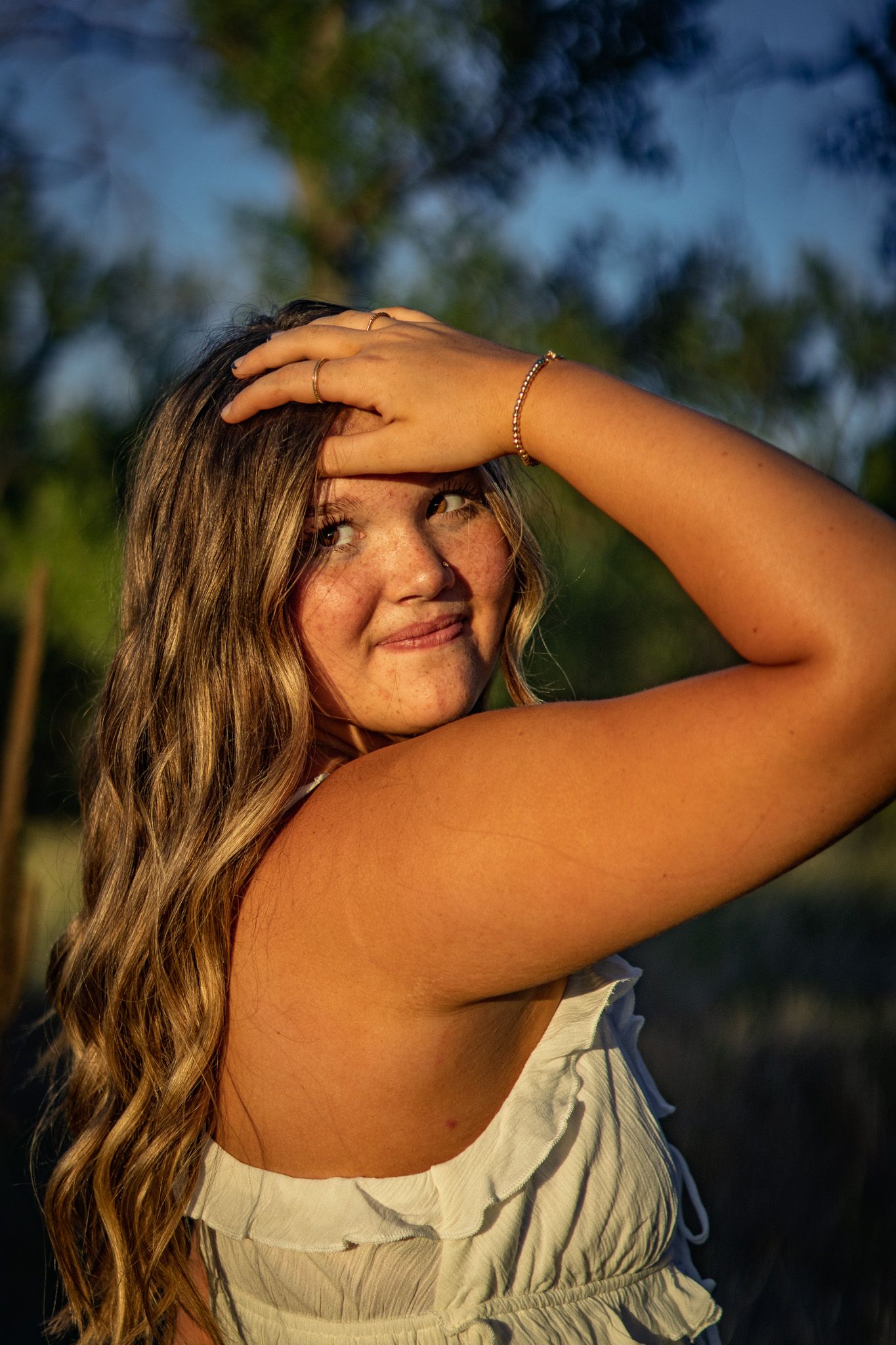 Young woman with long wavy hair shielding eyes from golden sunset light in outdoor portrait.