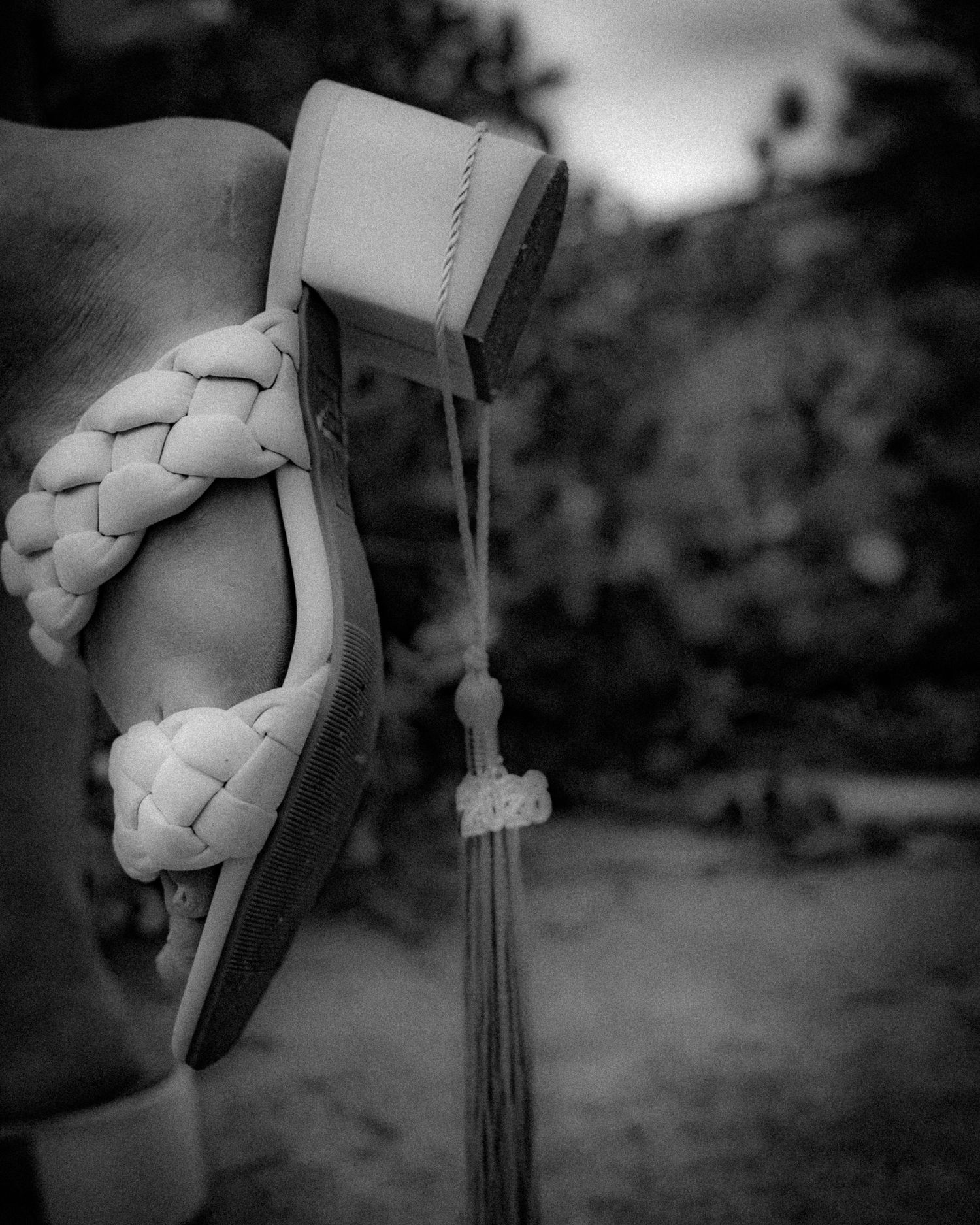 Close-up black and white photo of a braided sandal with tassel detail worn outdoors.