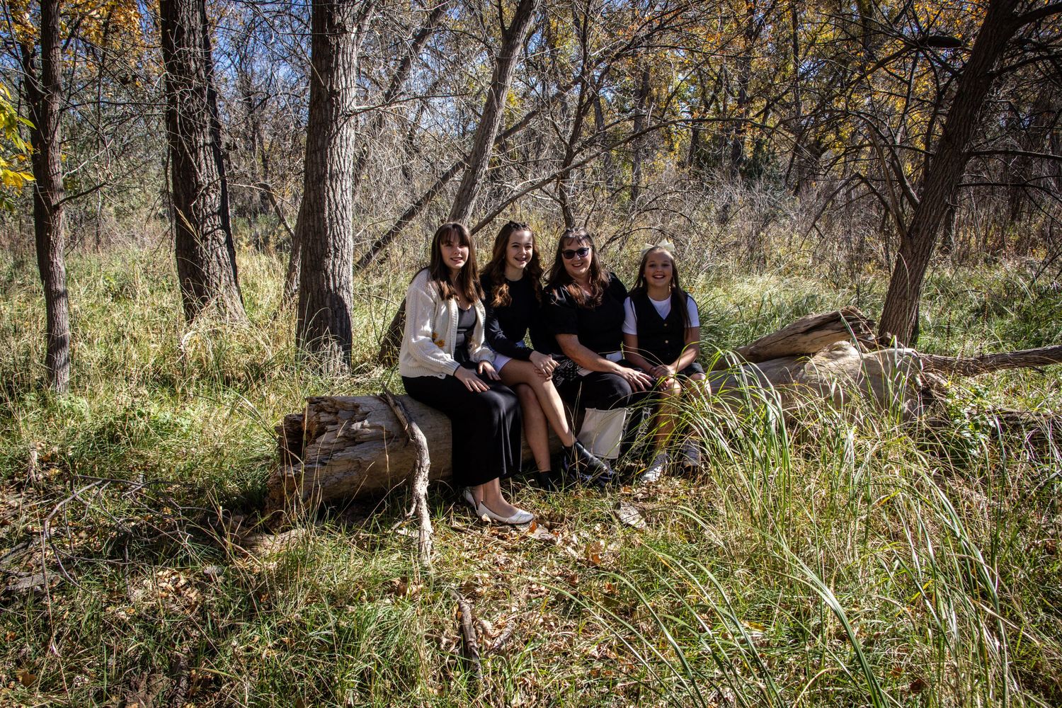 Four women sitting together on a log in a colorful autumn forest setting.