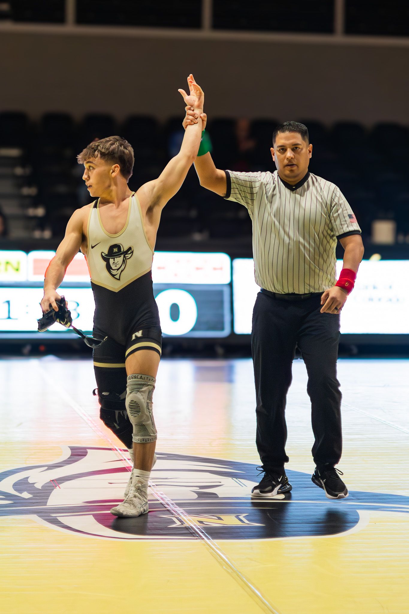 Wrestling referee raises the hand of a victorious athlete in a black singlet after a match win.