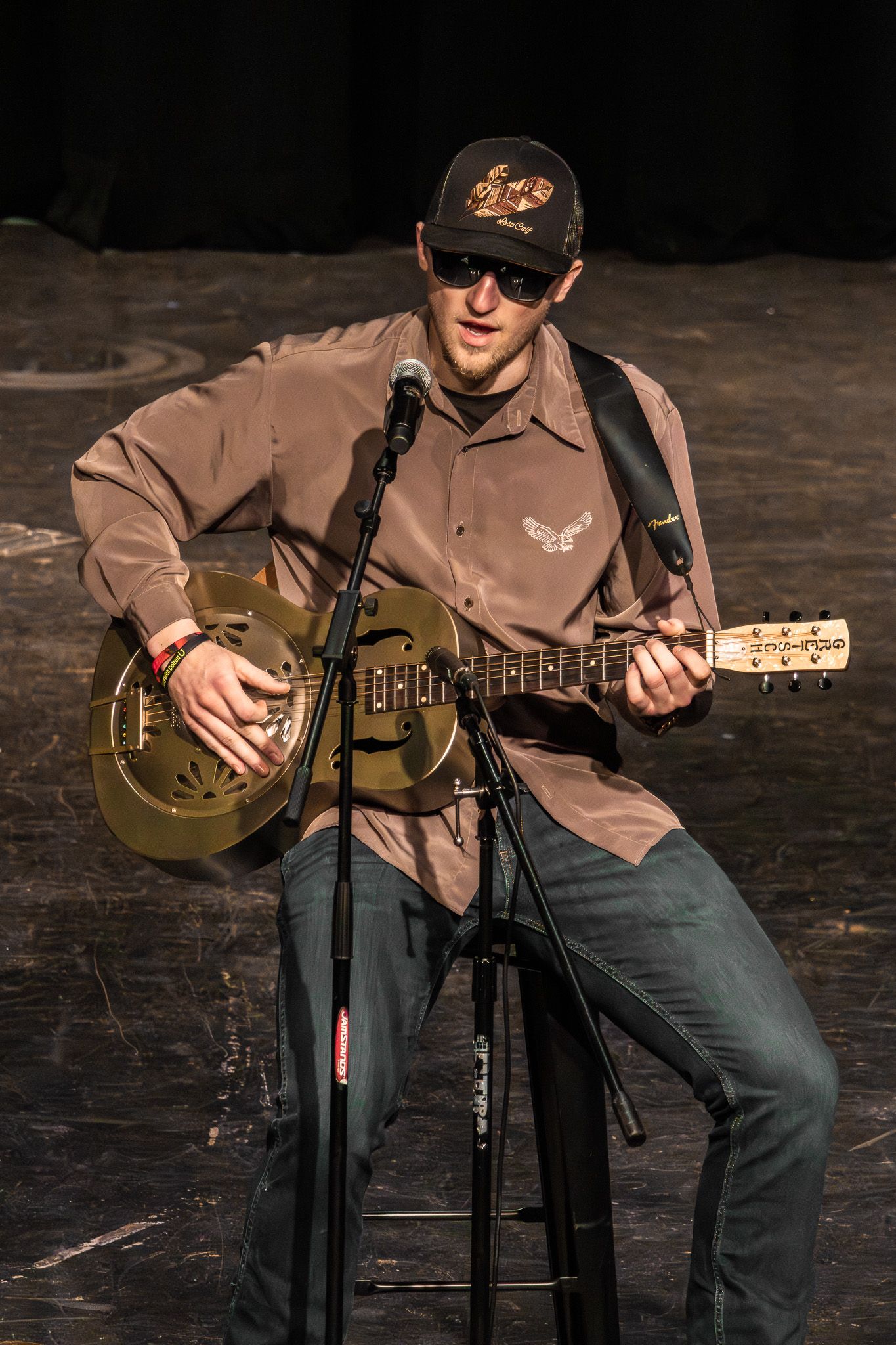 Male guitarist playing resonator guitar on stage wearing sunglasses and brown shirt at live music event.