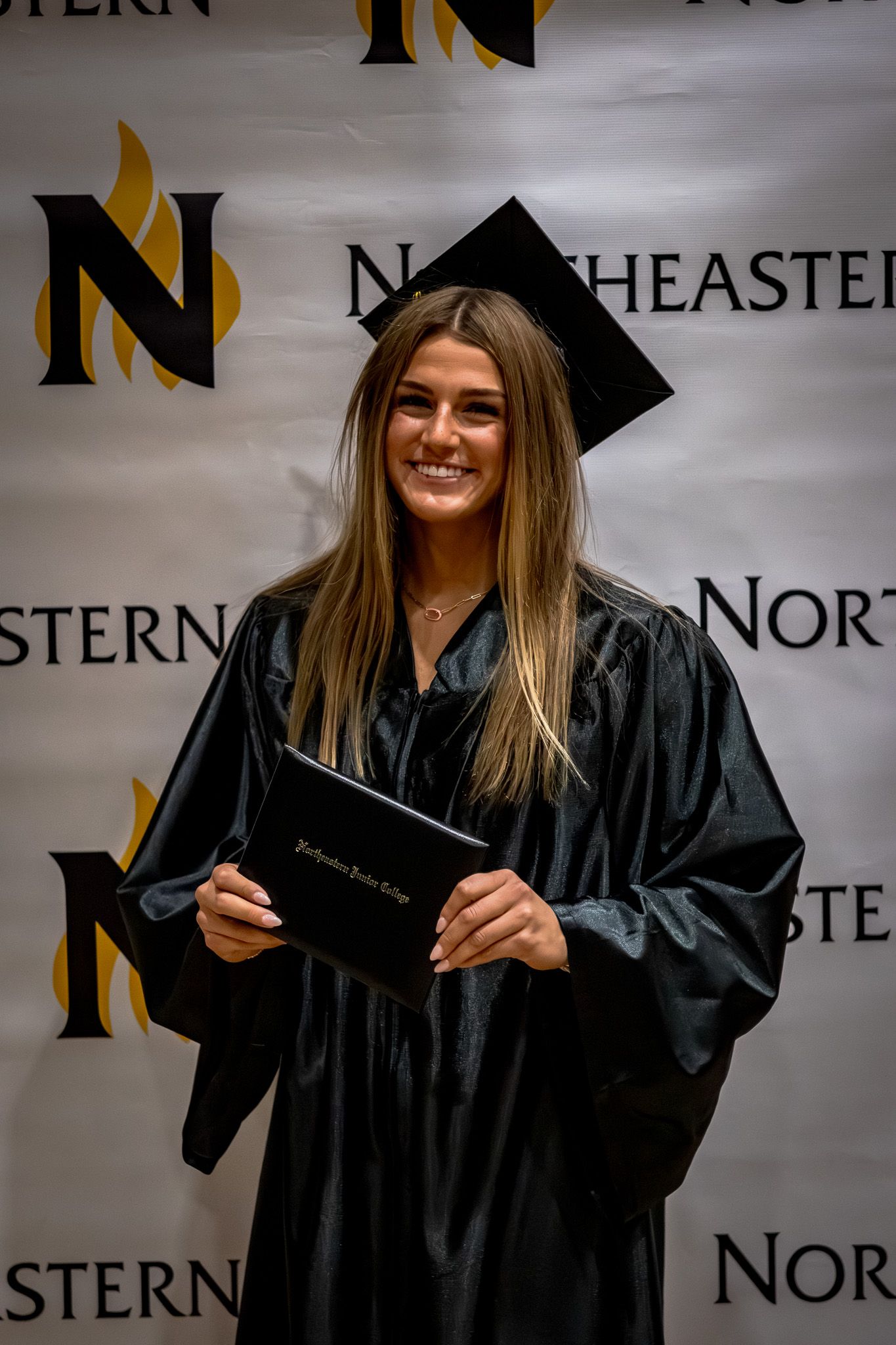 Smiling female graduate in black cap and gown holds diploma at Northeastern Junior College graduation backdrop.