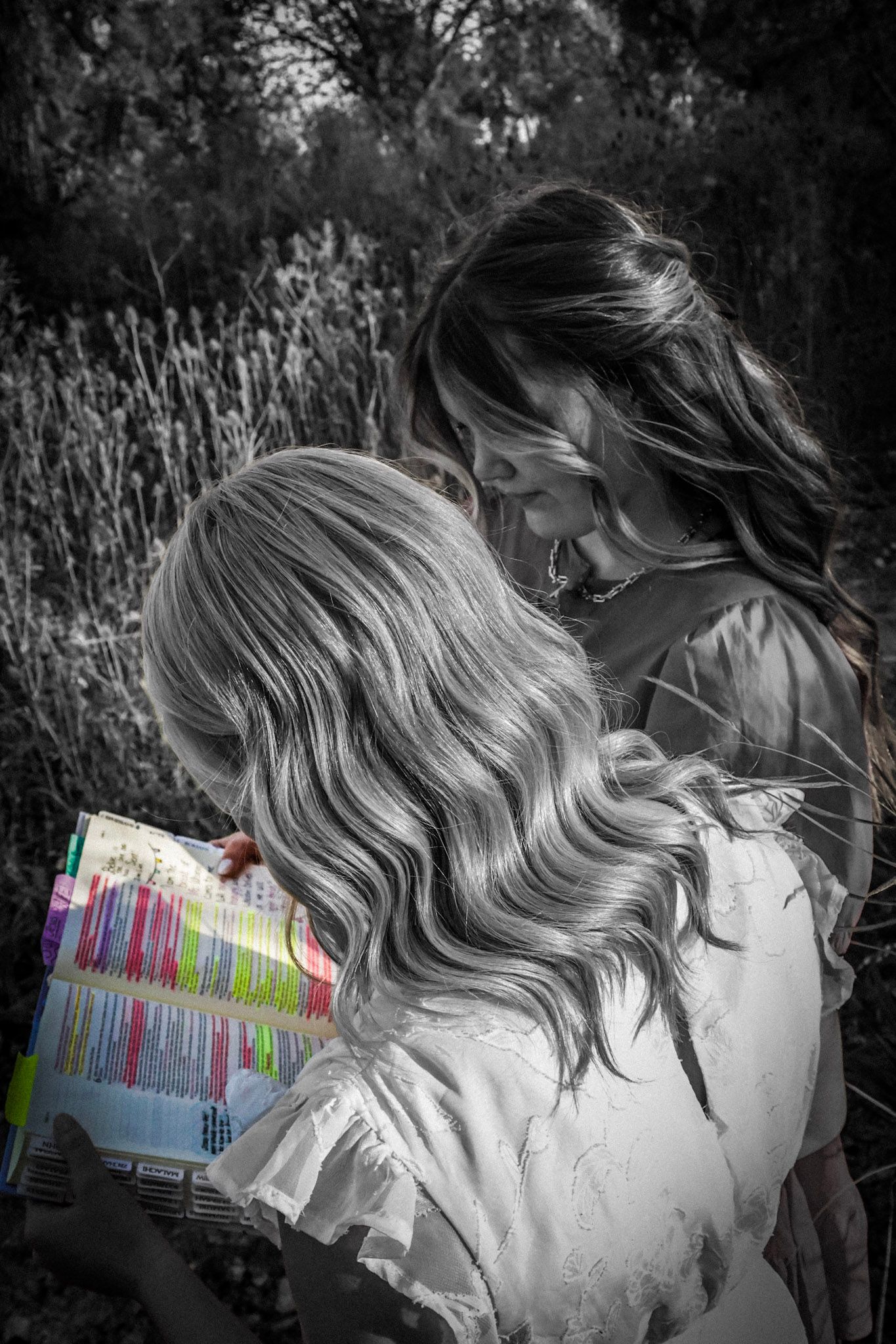 A black and white photo of two girls sharing a colorful book outdoors in a natural setting.