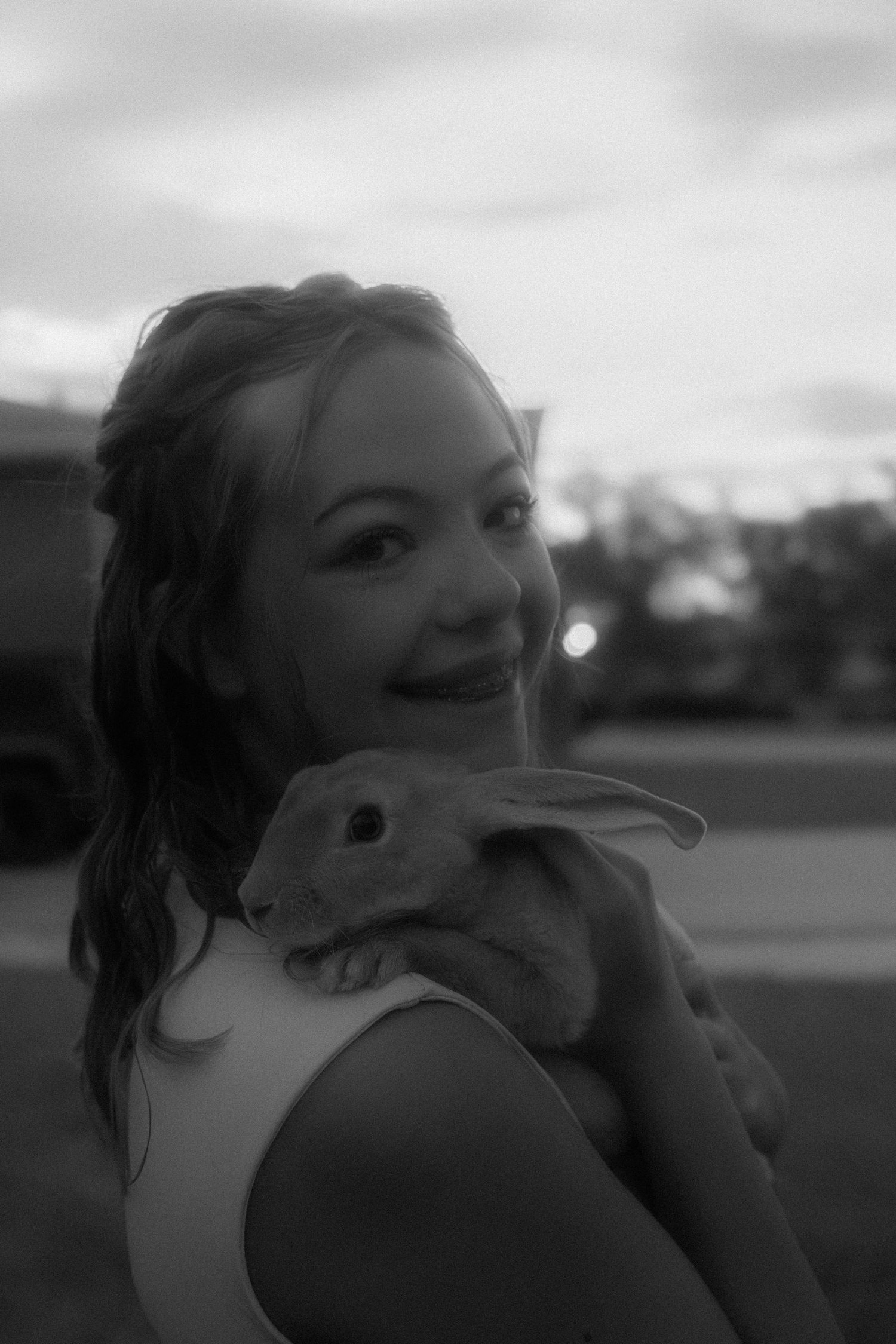 Black and white portrait of a smiling young girl holding a rabbit outdoors at dusk.