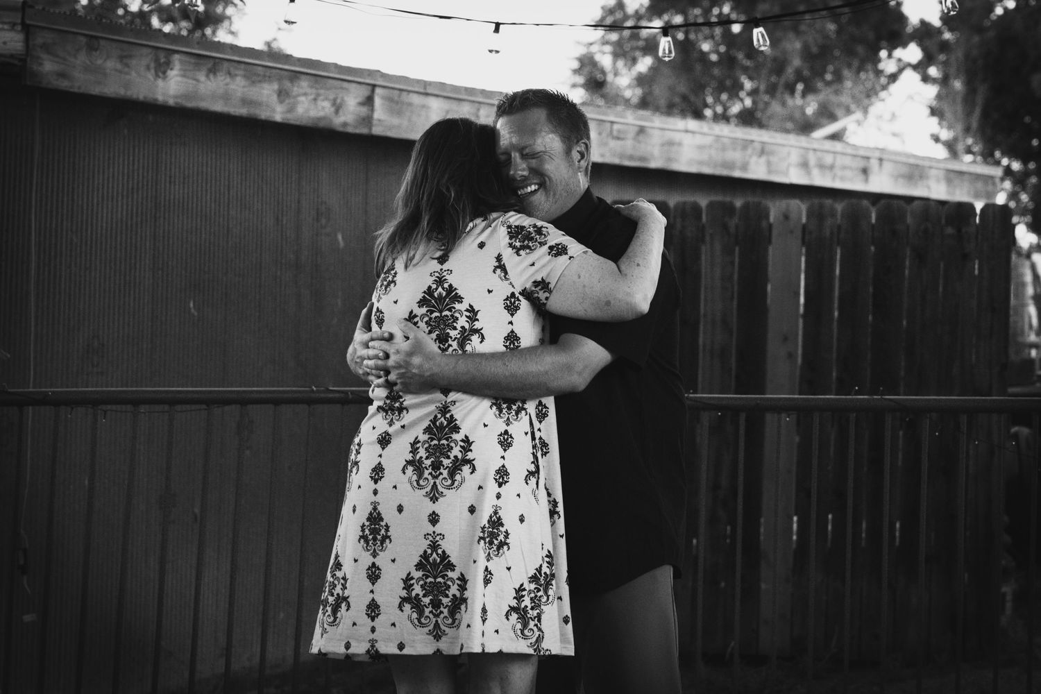 Black and white photo of a couple slow dancing and embracing in a backyard near a wooden fence.