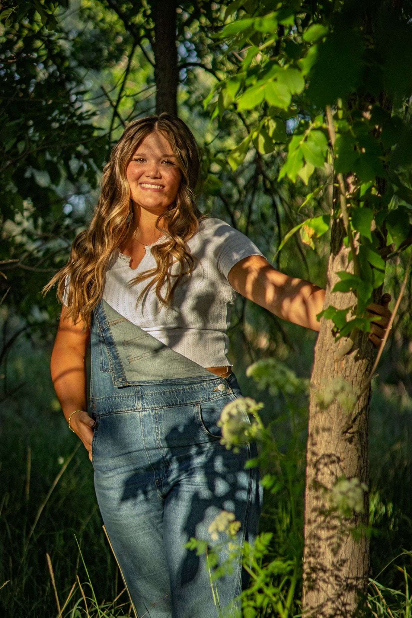 Smiling young woman in overalls leaning against a tree in dappled sunlight.