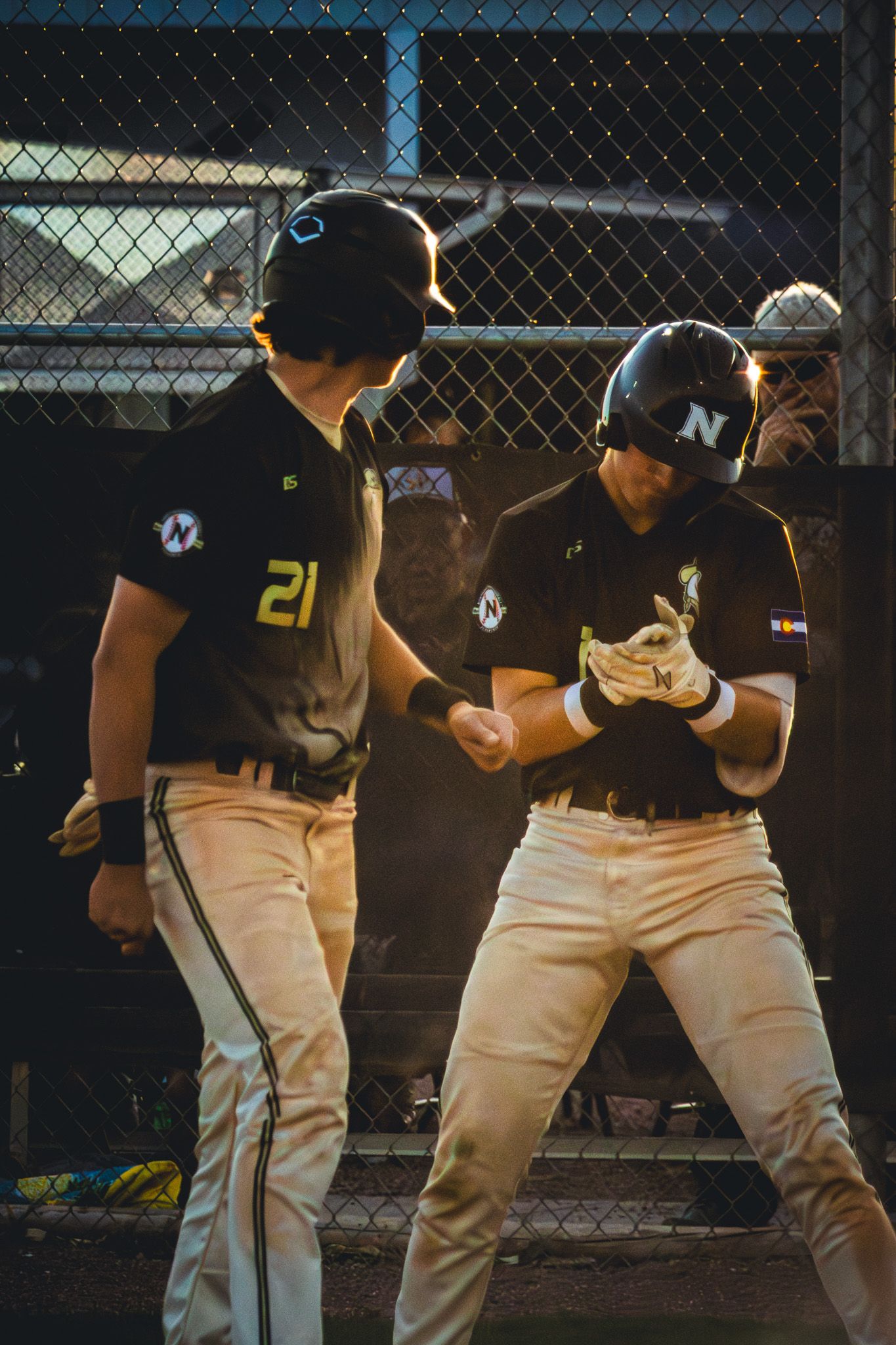 Two baseball players in black uniforms celebrate with a fist bump near a chain-link fence.