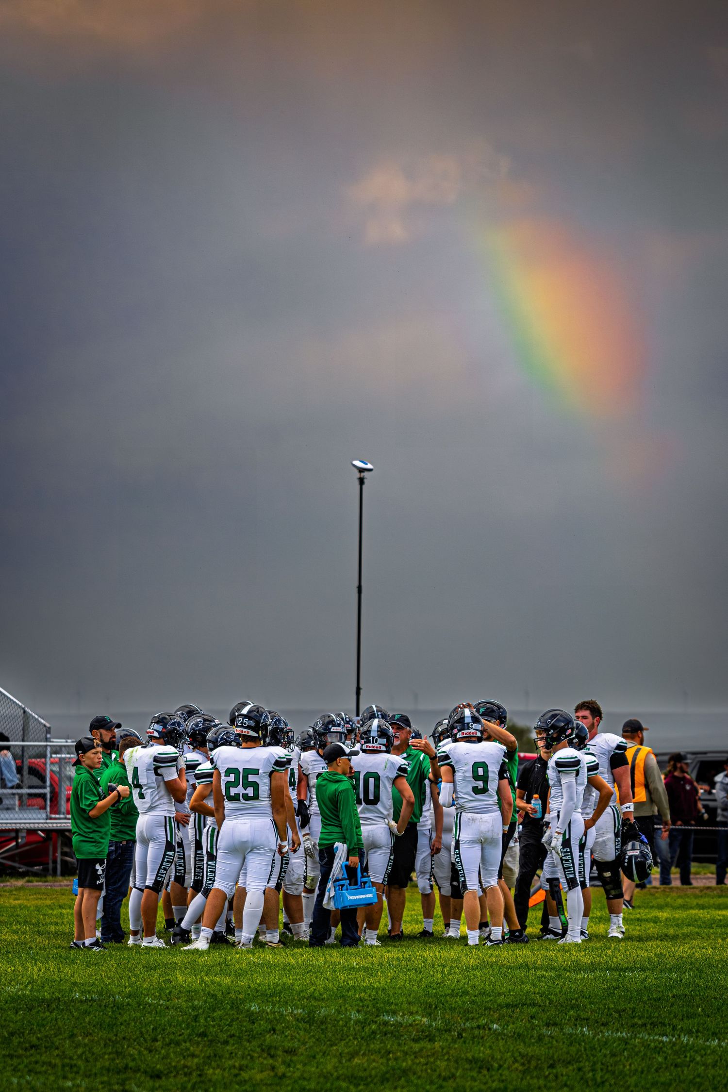 A football team huddles on the field as a rainbow appears through dramatic storm clouds above.