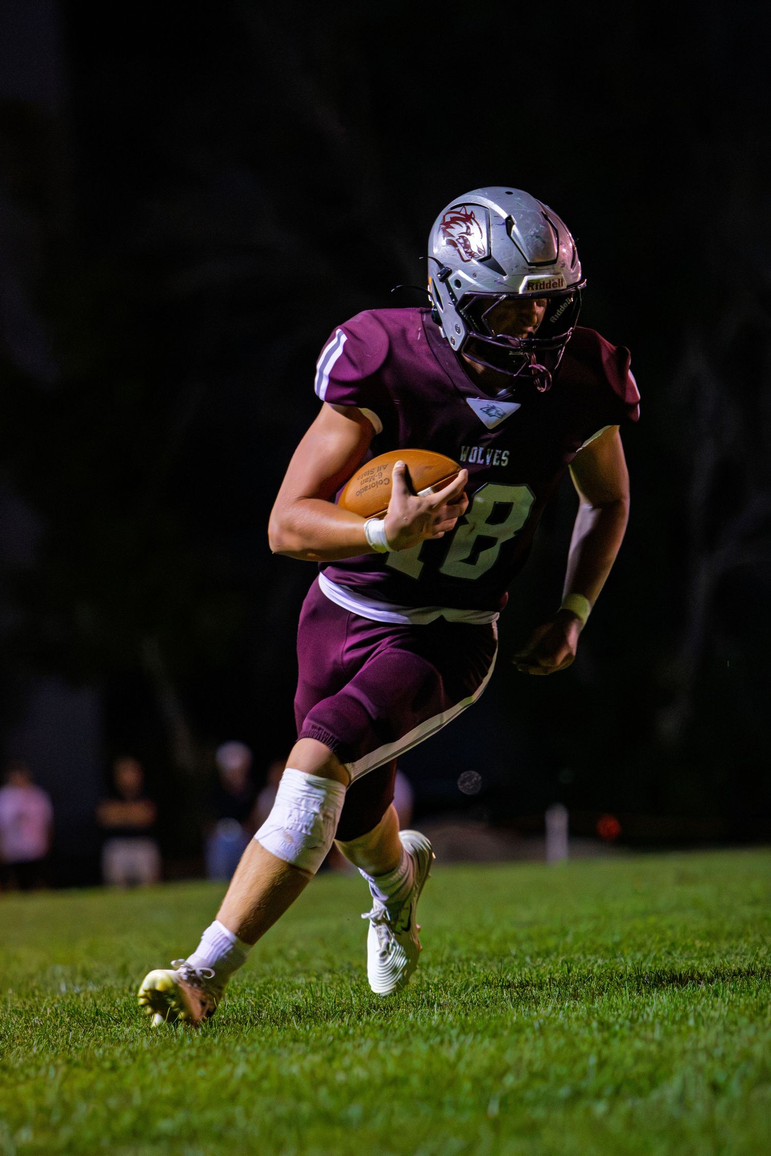 High school football player #18 in maroon uniform runs with the ball during a night game.
