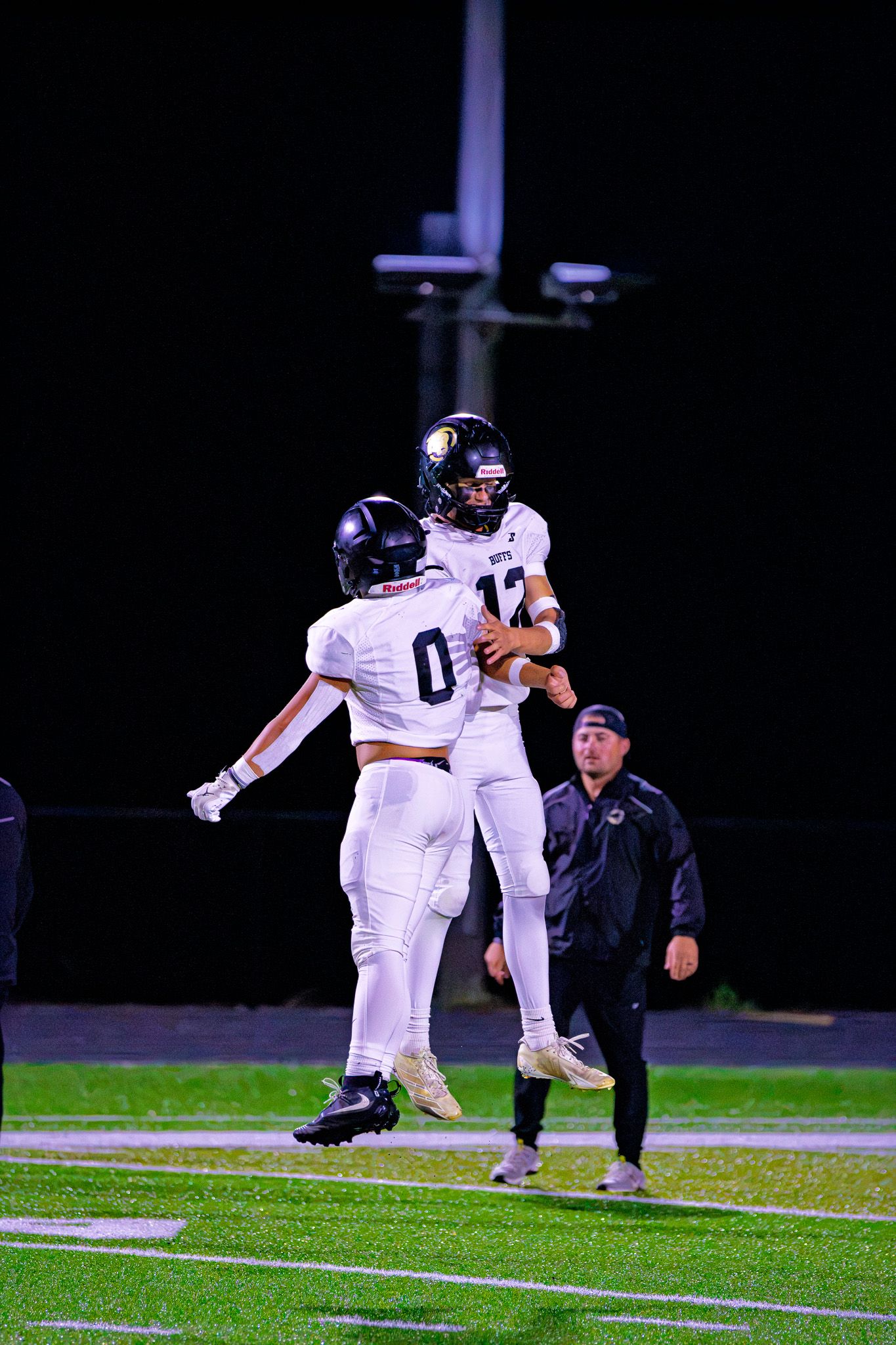 Two football players in white uniforms celebrate a touchdown with a chest bump on the field at night.