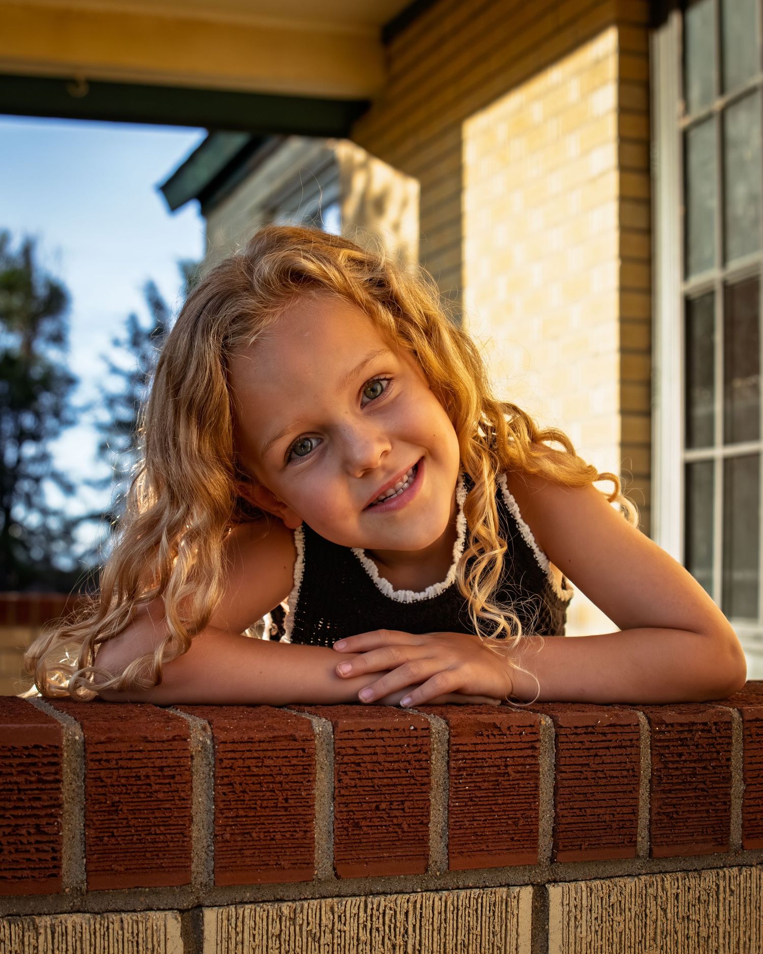 Adorable young girl with curly blonde hair smiles while leaning on a brick wall outdoors.