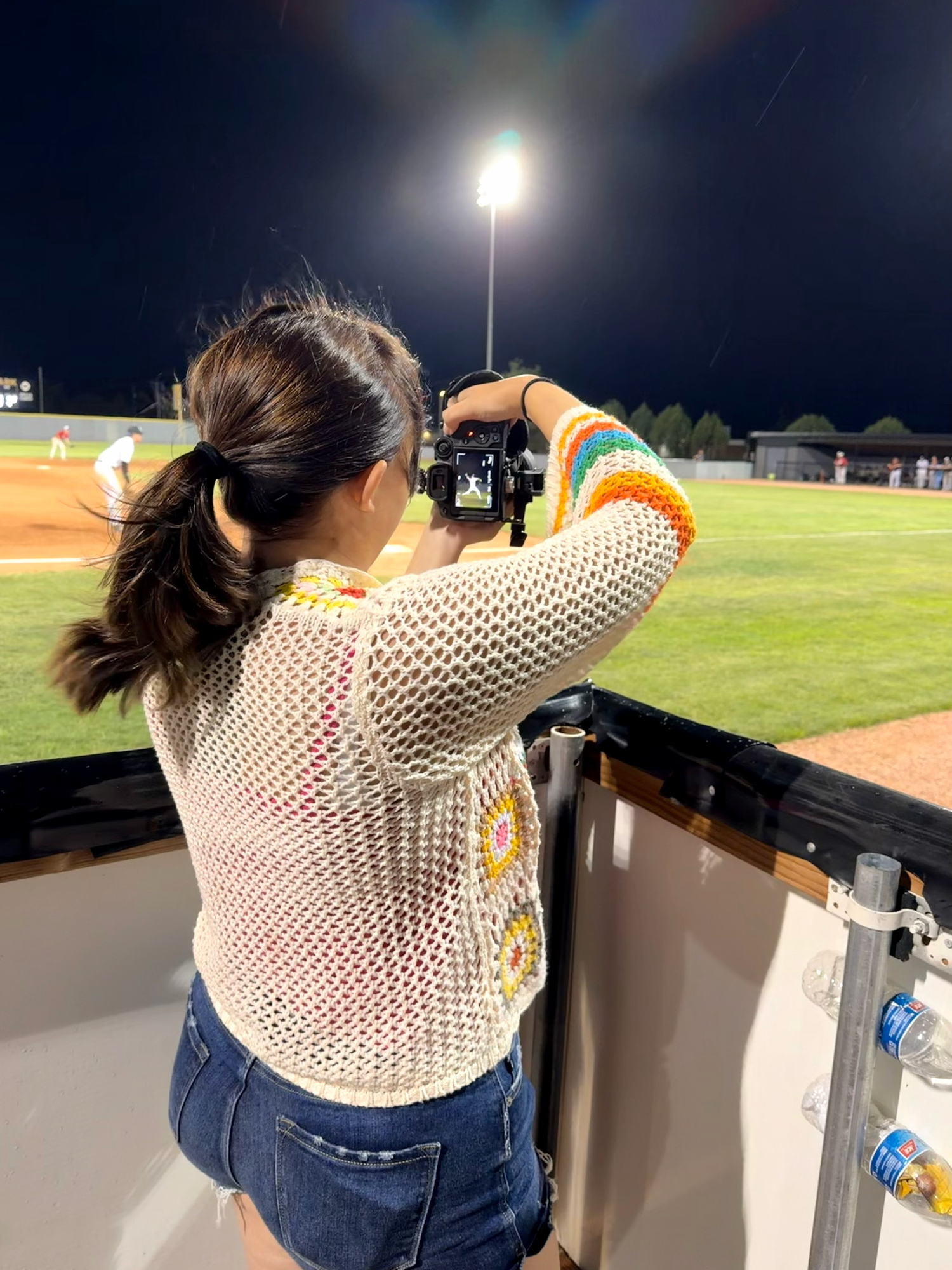 Woman in crochet sweater photographs a night baseball game from the dugout with a DSLR camera.