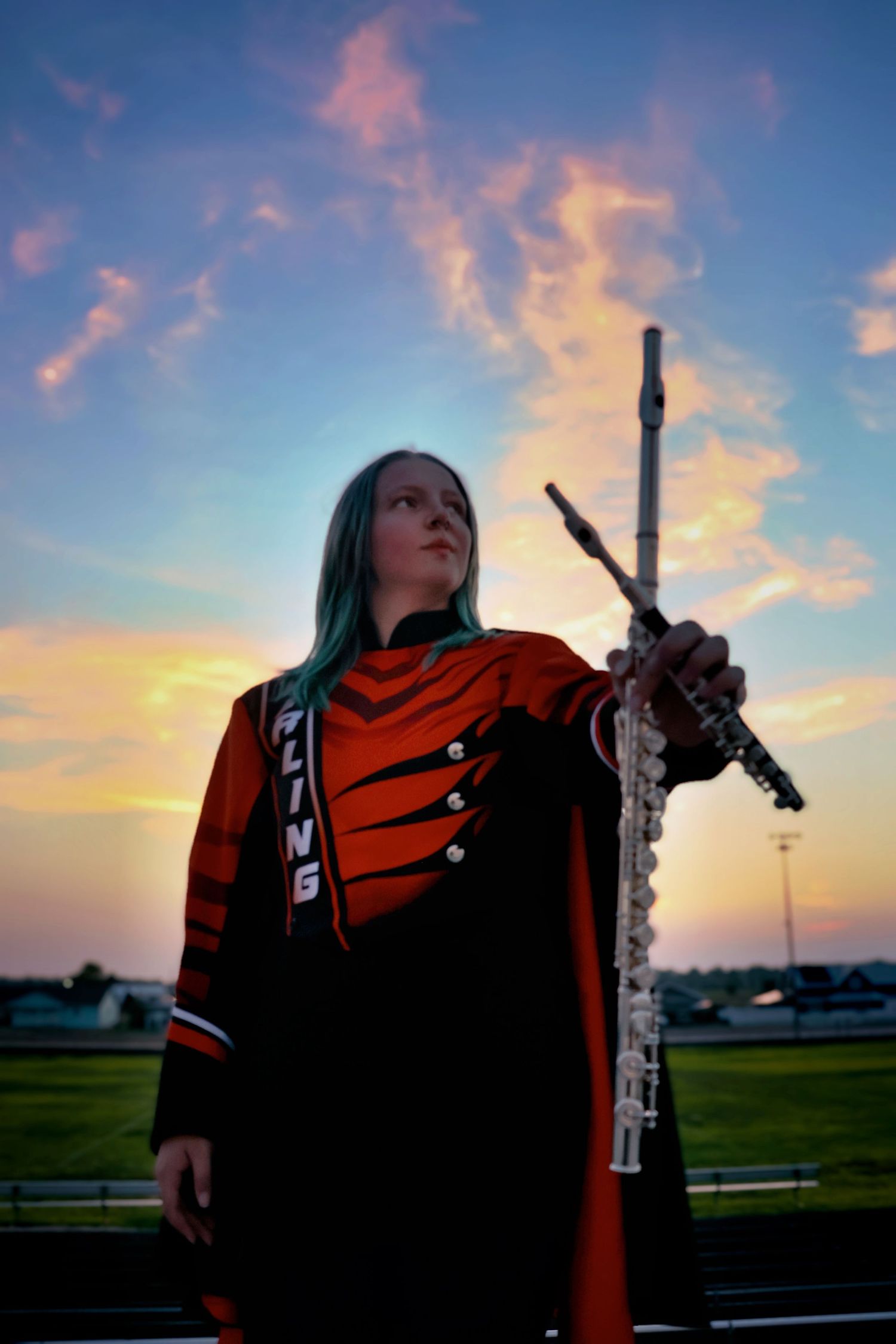 Female marching band flutist in orange uniform holds flute against a vibrant colorful sunset sky.