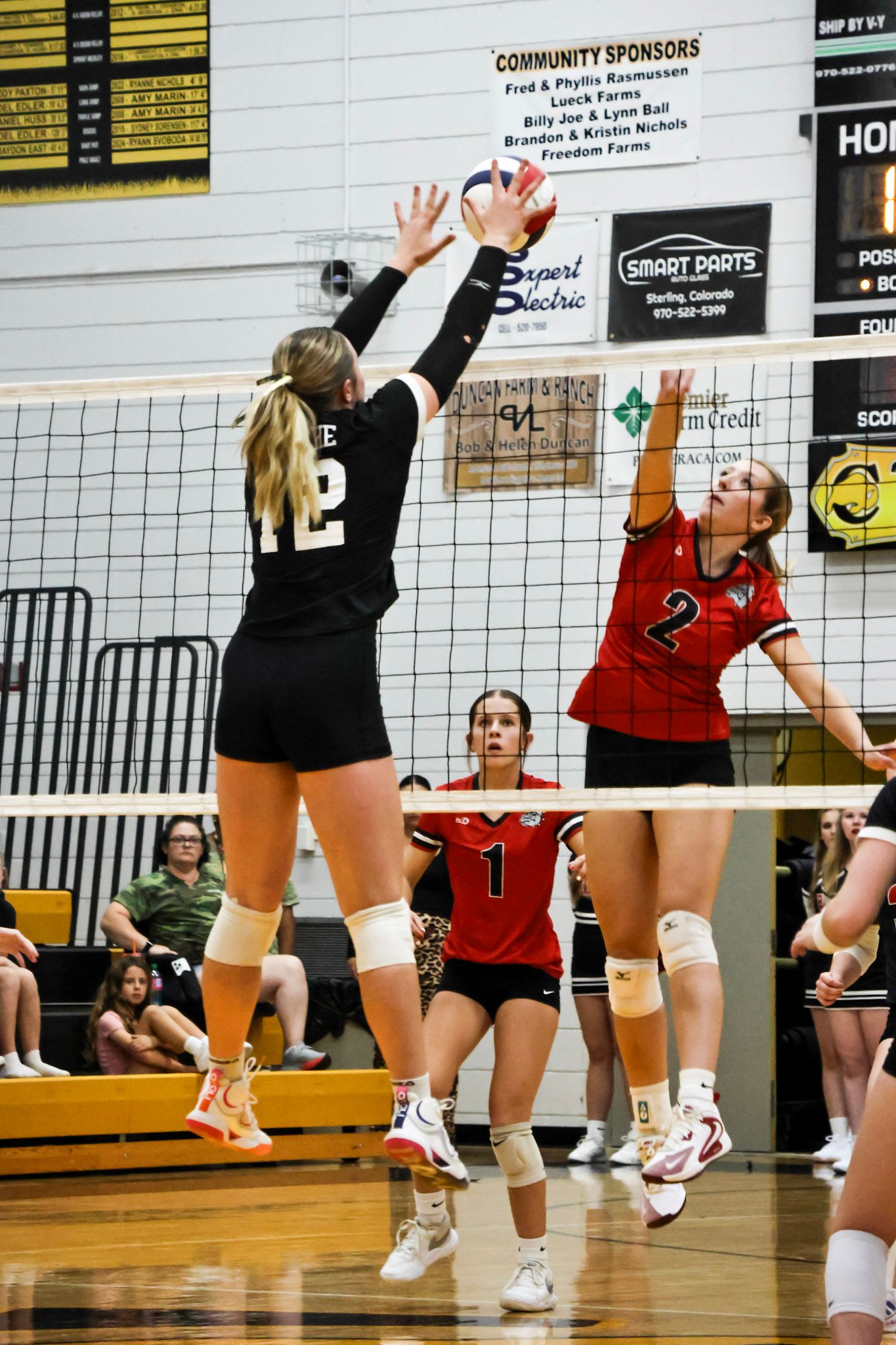 Volleyball player #12 blocking at the net against opponent #2 during an indoor match.